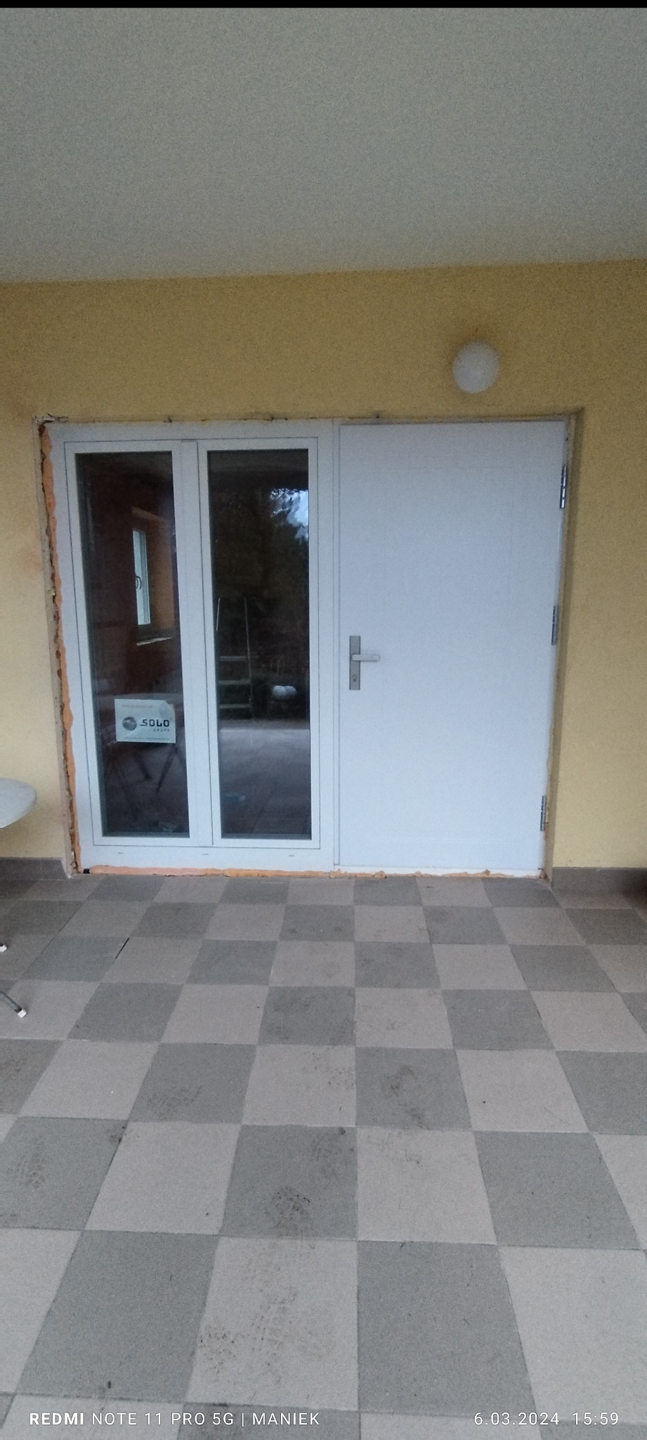 Białe drzwi wejściowe z szybą, zamontowane w żółtej ścianie. Widoczne ślady pianki montażowej. Na szybie naklejka informacyjna. Podłoga wyłożona szarą szachownicą.