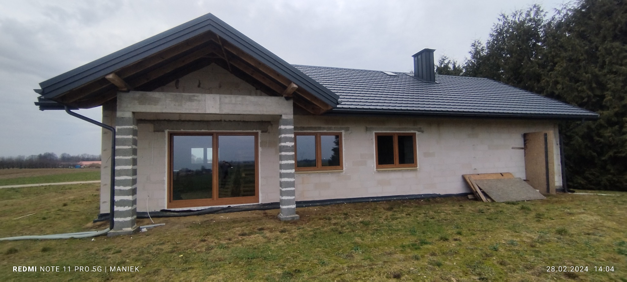 Dom w trakcie budowy z zamontowanymi oknami i drzwiami z brązowymi ramami, widoczne bloczki betonowe i szara dachówka. Krajobraz wiejski w tle.