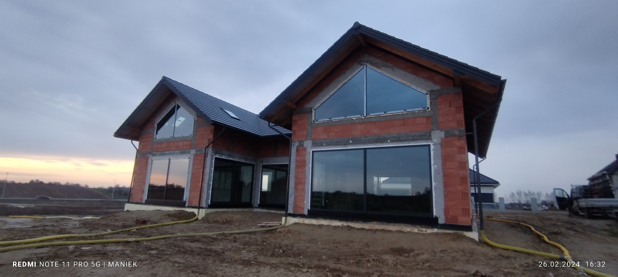 Dom w stanie surowym otwartym z zamontowanymi oknami w Turce. Widoczne duże przeszklenia i ceglana konstrukcja. Dach pokryty dachówką. Krajobraz w tle.