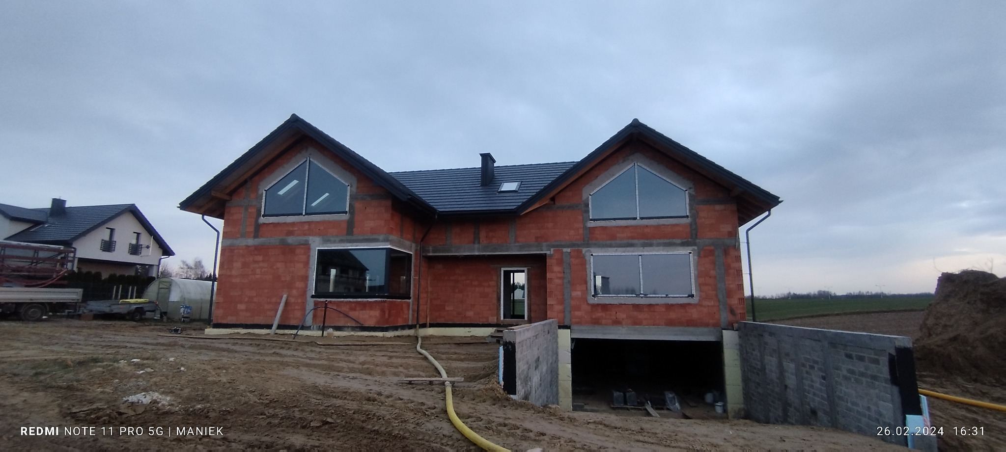 Surowy stan domu z czerwonej cegły z zamontowanymi oknami o nietypowych kształtach, widok z zewnątrz na tle pochmurnego nieba i ziemi.