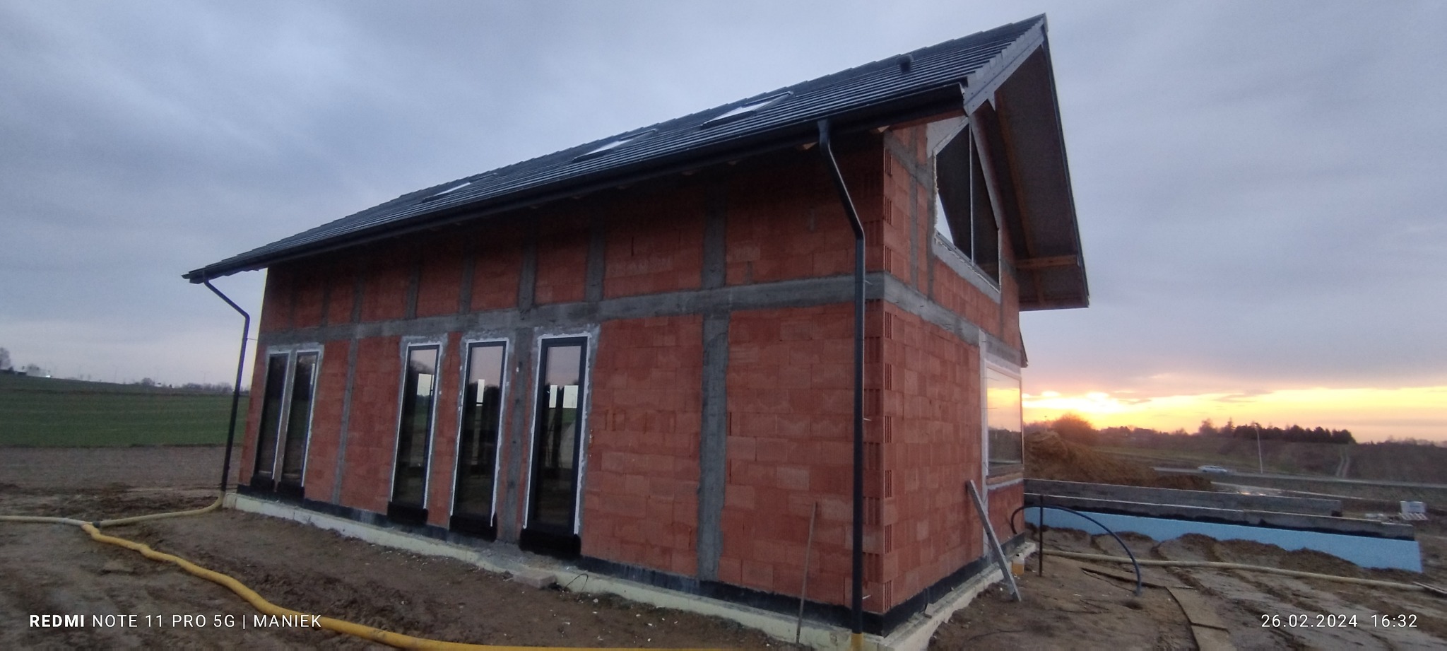 Surowy budynek z czerwonej cegły z zamontowanymi oknami PCV, widok z zewnątrz o zmierzchu. Widoczne elementy konstrukcyjne i instalacyjne.