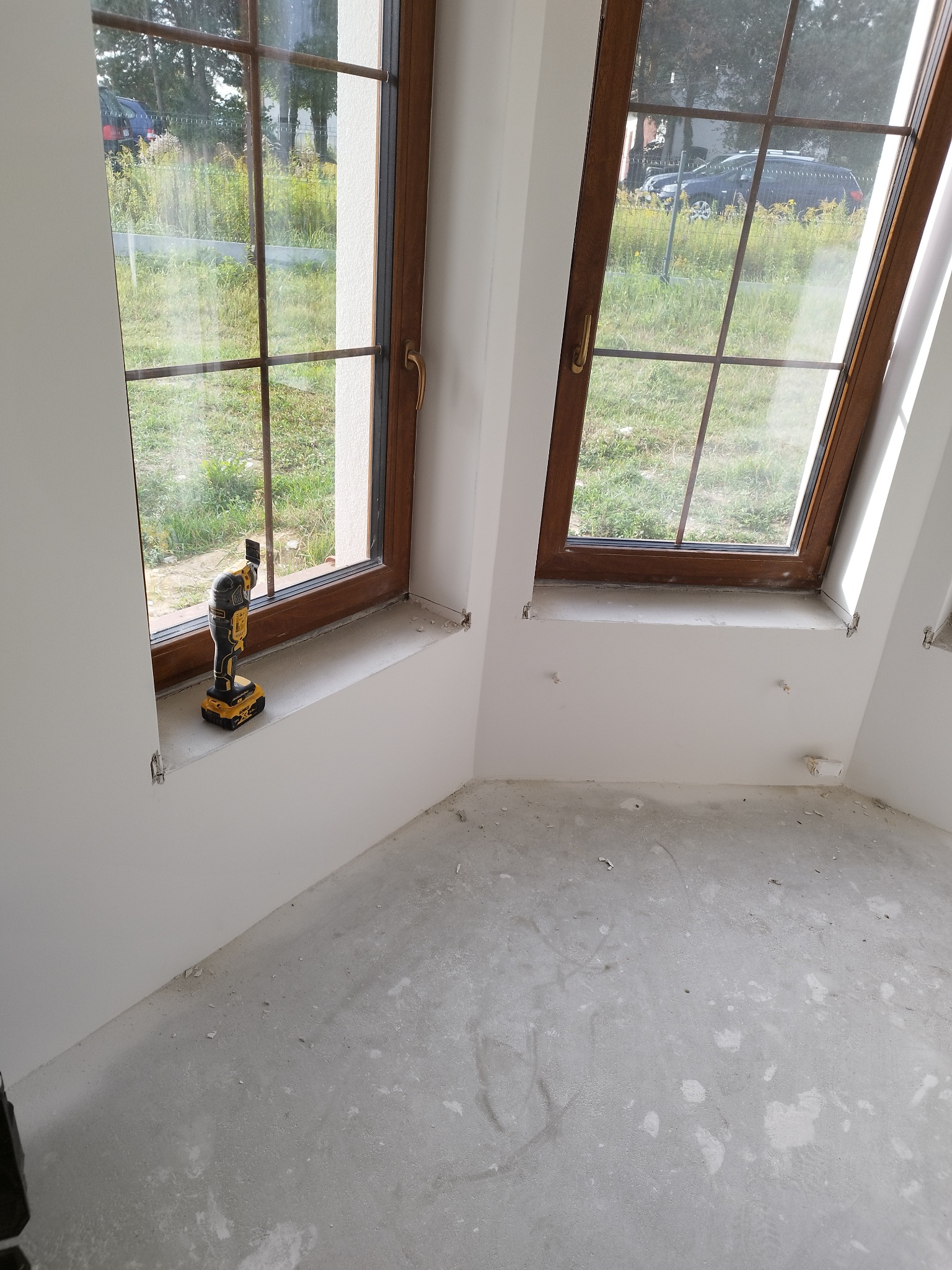 Montaż okien drewnianych z brązowymi ramami w nowym budynku. Na parapecie stoi narzędzie wielofunkcyjne DeWalt. Widok na zewnątrz przez okno.