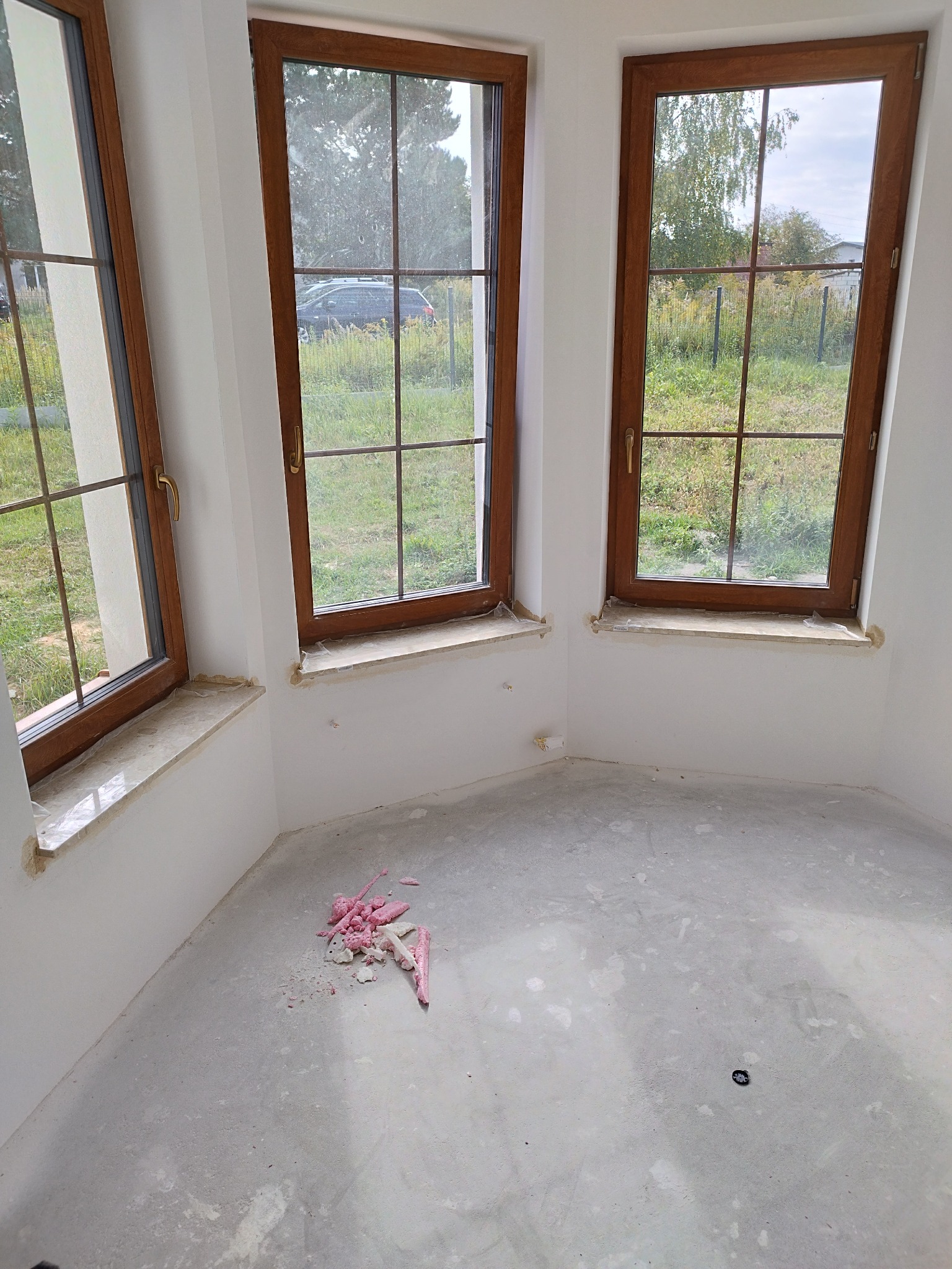 Wnętrze z trzema oknami z brązowymi ramami i szprosami, widok na zewnątrz, na podłodze resztki pianki montażowej, ściany białe, surowy stan wykończenia.