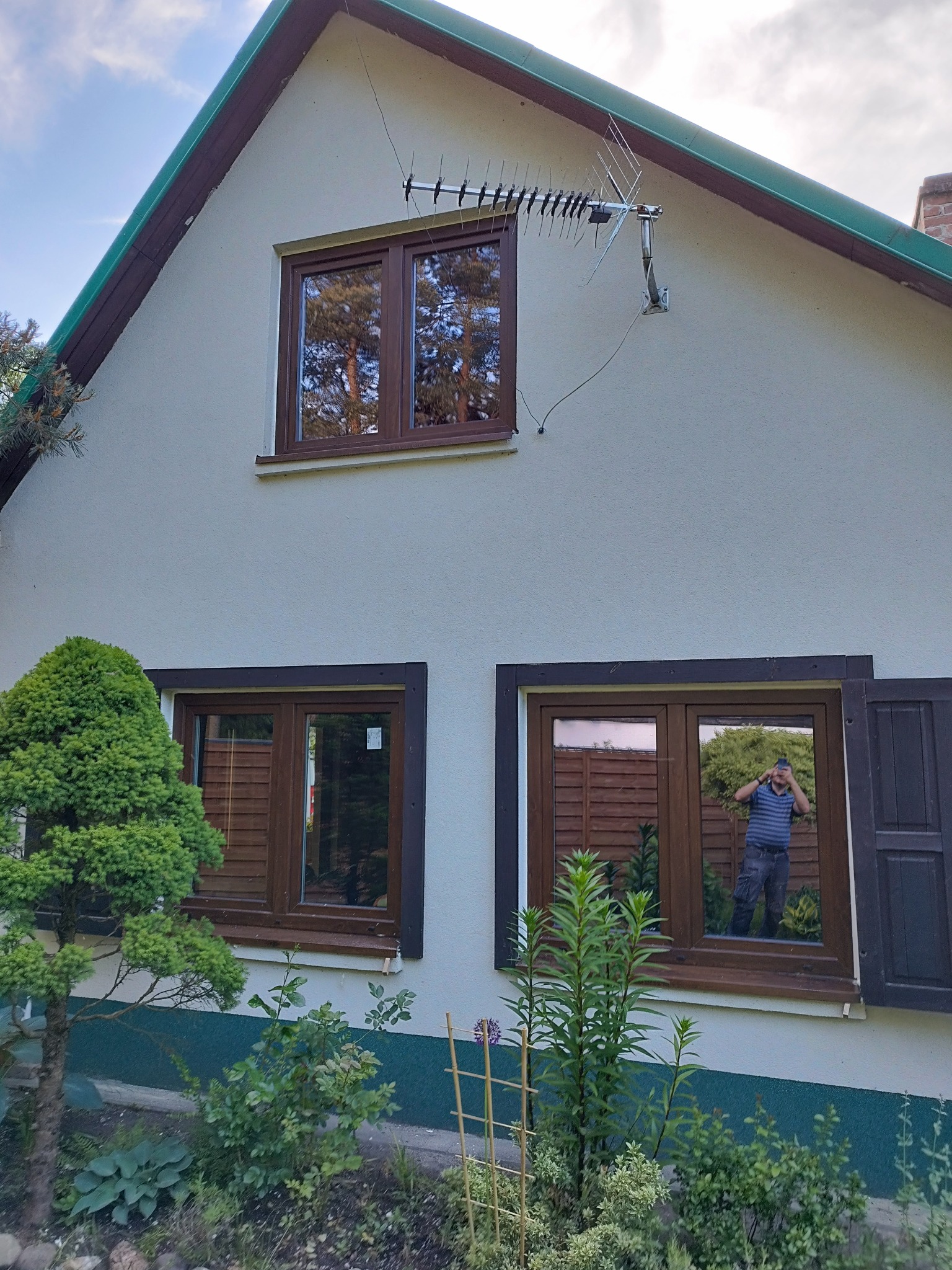 Dom z brązowymi oknami, w których odbija się otoczenie i osoba robiąca zdjęcie. Na ścianie antena. Otwarta okiennica po prawej stronie.