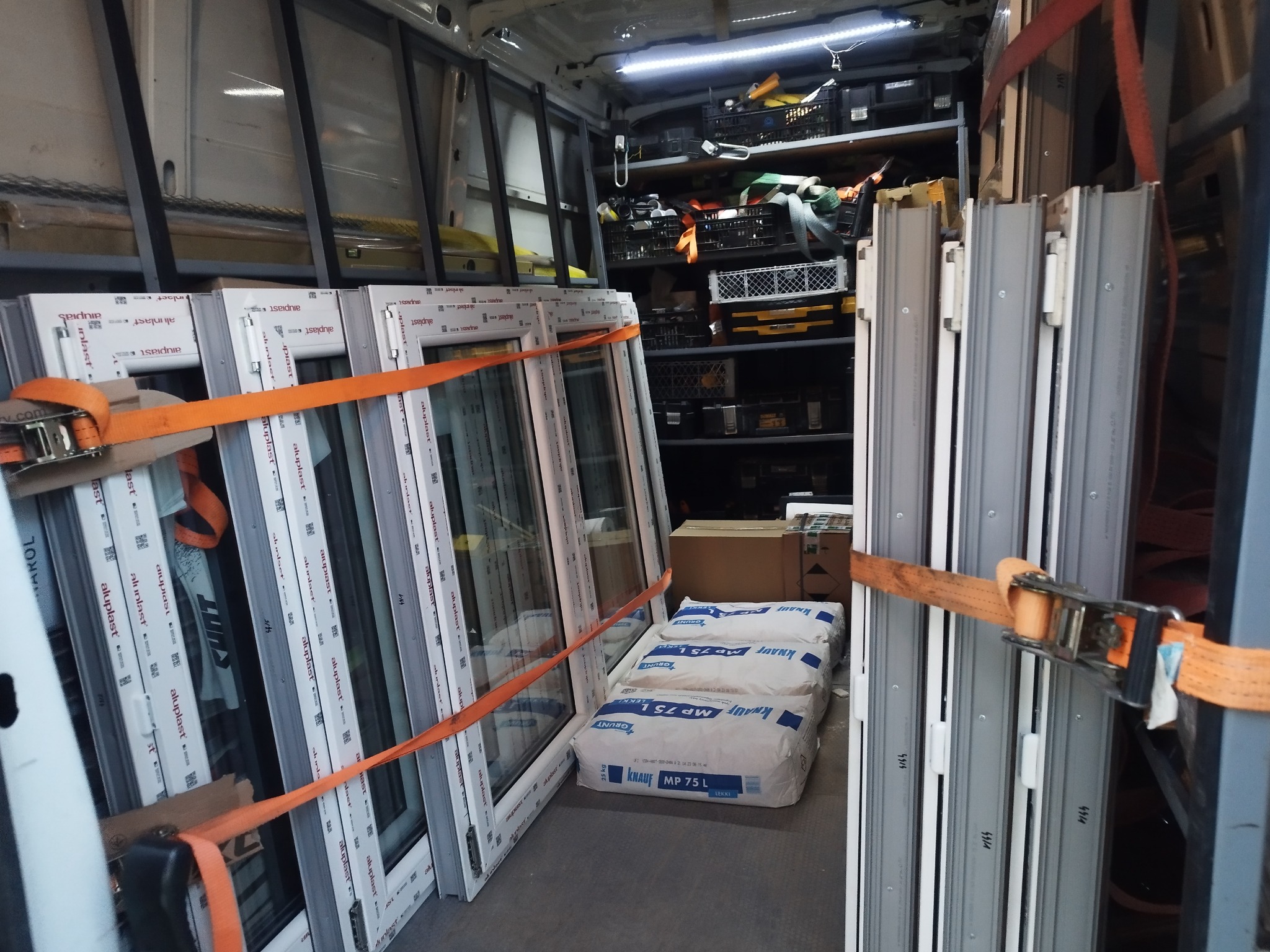 Transport okien PCV w busie, zabezpieczonych pomarańczowymi pasami. Widoczne worki z zaprawą i narzędzia na półkach. Ujęcie z wnętrza pojazdu.