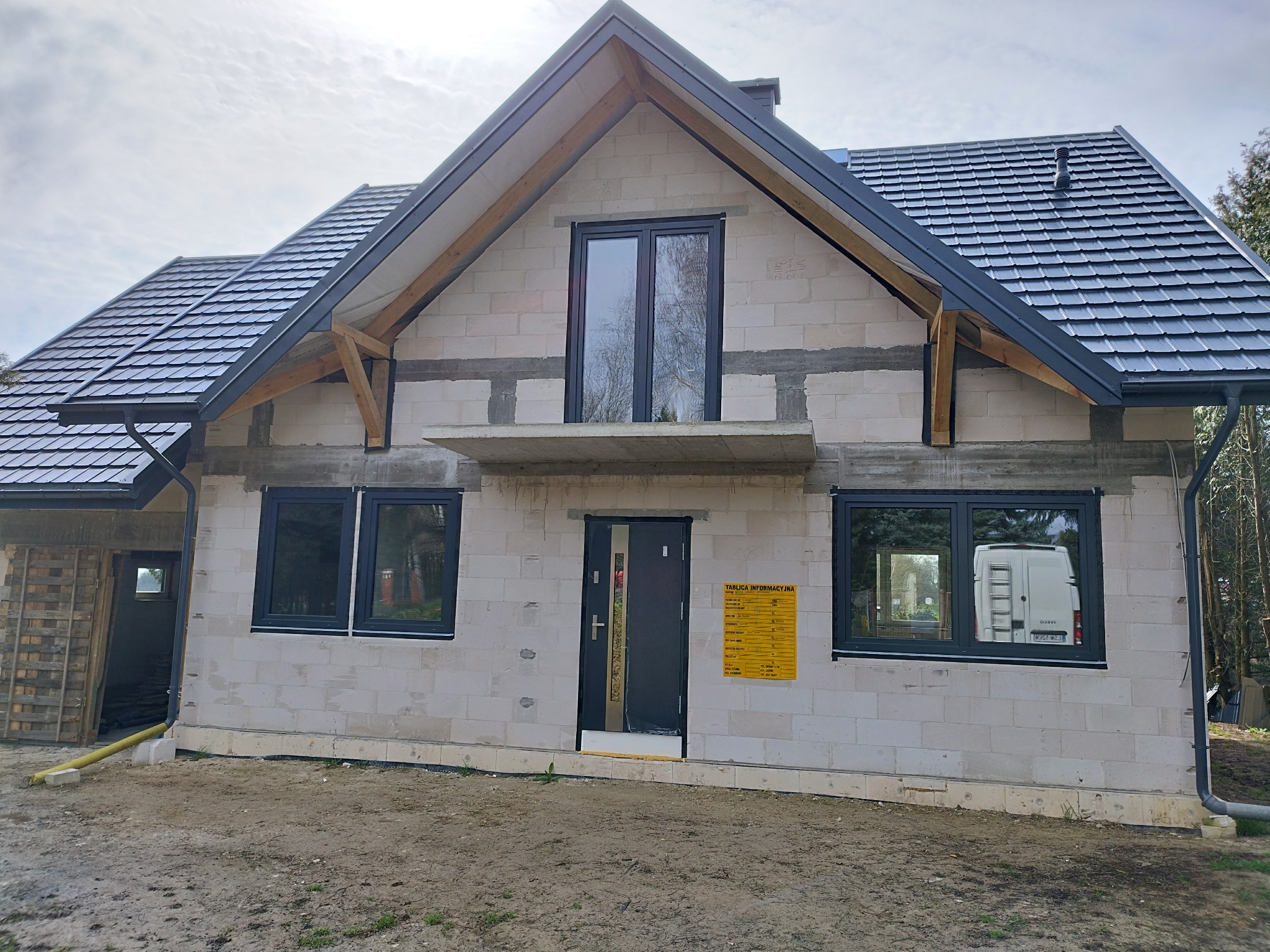 Dom w trakcie budowy z zamontowanymi oknami w grafitowej ramie i ciemnymi drzwiami wejściowymi. Na ścianie wisi żółta tablica informacyjna.