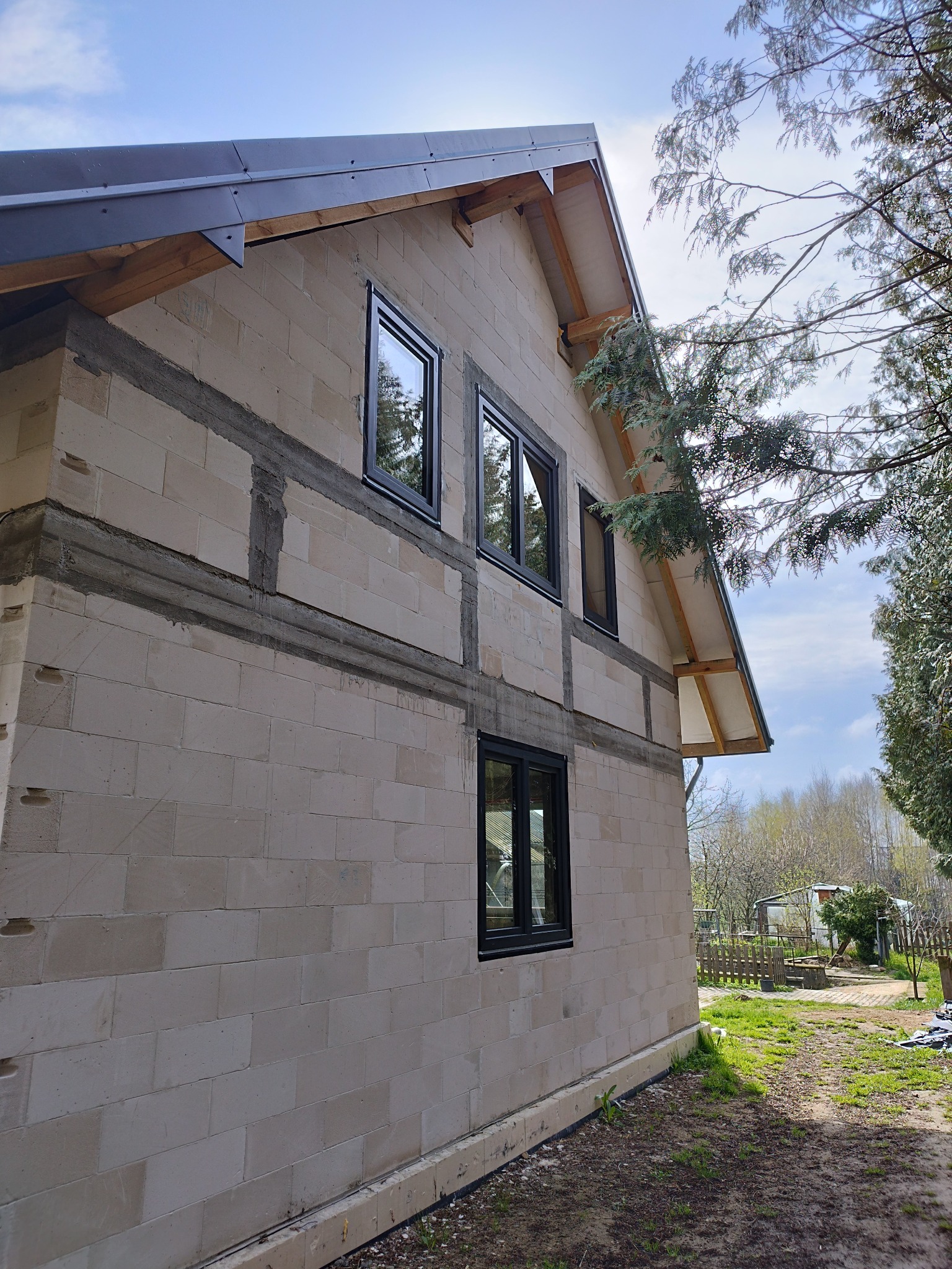 Surowy stan budynku z czarnymi oknami, widoczne bloczki i betonowe wzmocnienia, drewniana konstrukcja dachu, otoczenie zieleni i nieba.