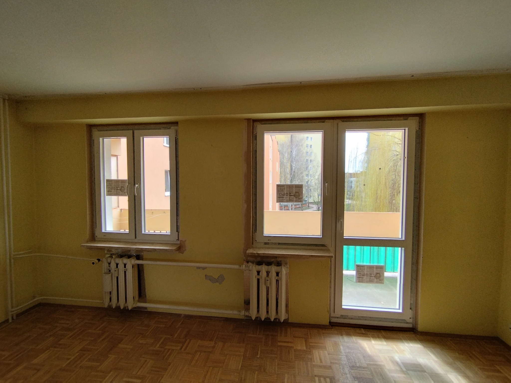 Wnętrze pokoju z dwoma oknami PCV i grzejnikami. Widoczne ślady po remoncie na ścianach i podłodze. Jedno z okien pełni funkcję drzwi balkonowych.