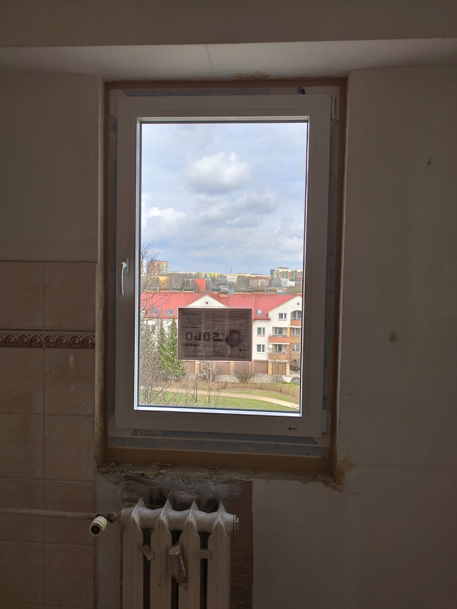Białe okno PCV w trakcie montażu, widok na budynki mieszkalne i pochmurne niebo. Grzejnik pod oknem, ślady po remoncie na ścianach.