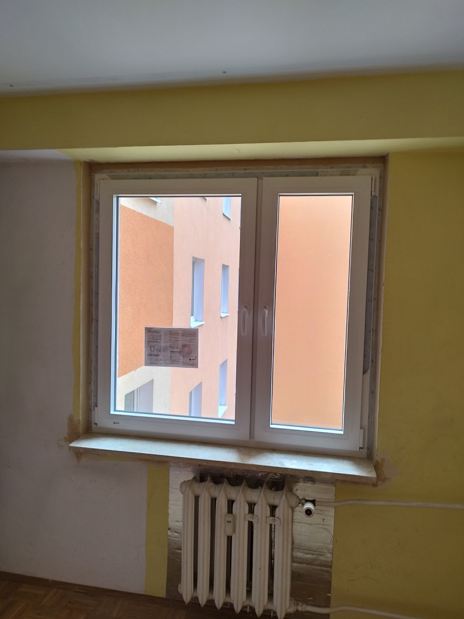 Białe okno PCV z widokiem na budynek w odcieniach pomarańczy, zamontowane w pokoju z żółtymi ścianami i grzejnikiem pod parapetem. Widoczne ślady po remoncie.