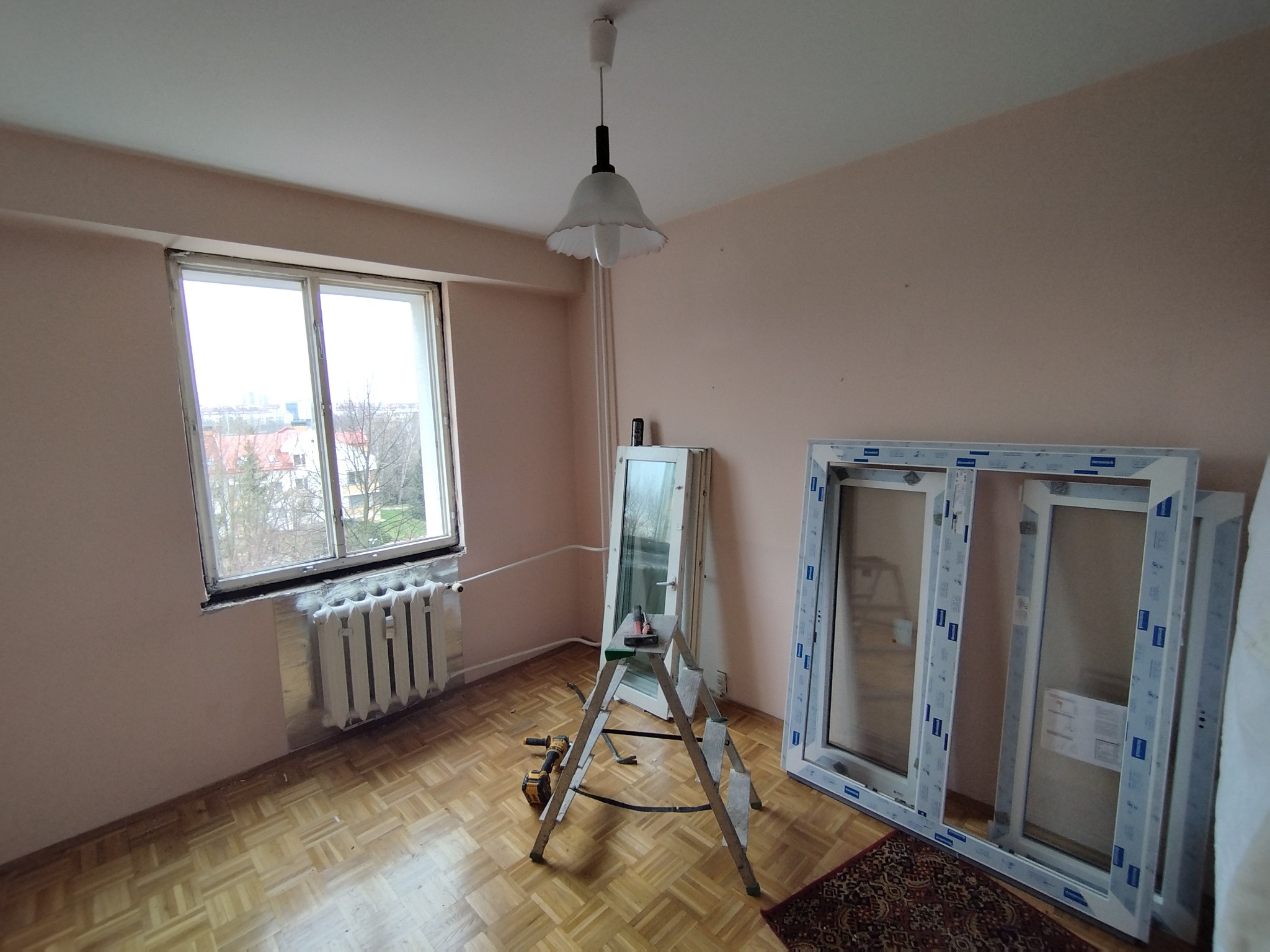 Pokój w trakcie wymiany okna: stare okno usunięte, nowe okna PCV oparte o ścianę, drabina z narzędziami, parkiet, grzejnik. Widok z okna na drzewa i budynki.