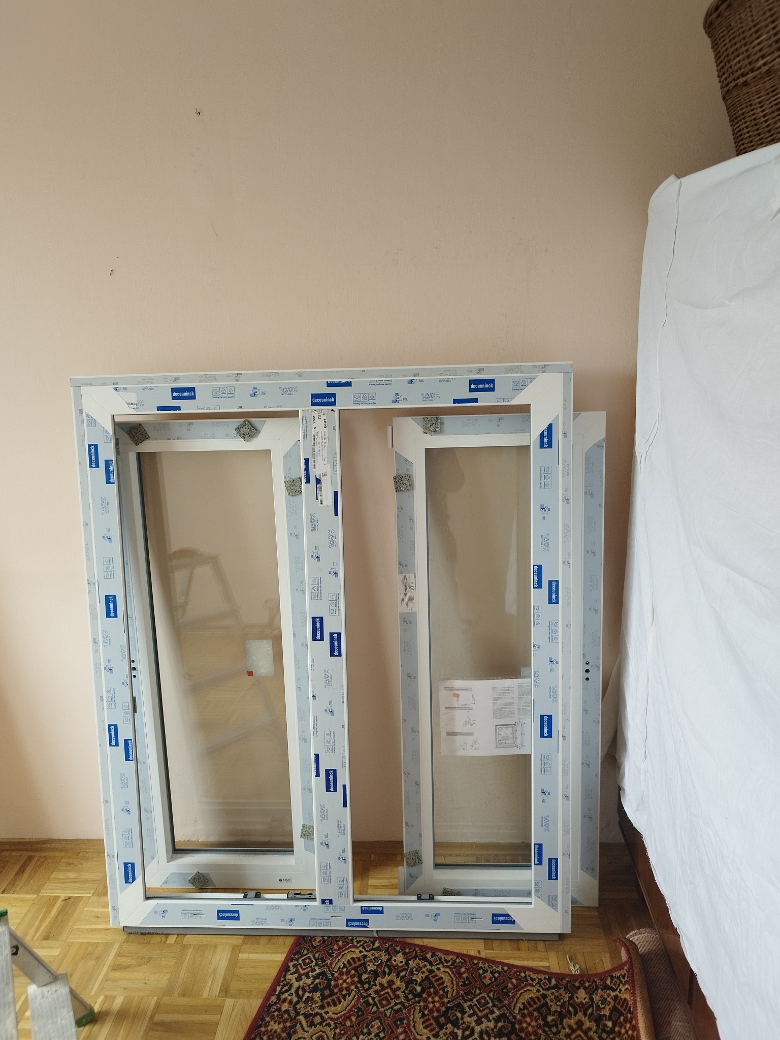 Dwa białe ramy okienne PCV Deceuninck, zabezpieczone folią, oparte o ścianę w pomieszczeniu z parkietem i dywanem. Widoczna drabina i fragment łóżka.
