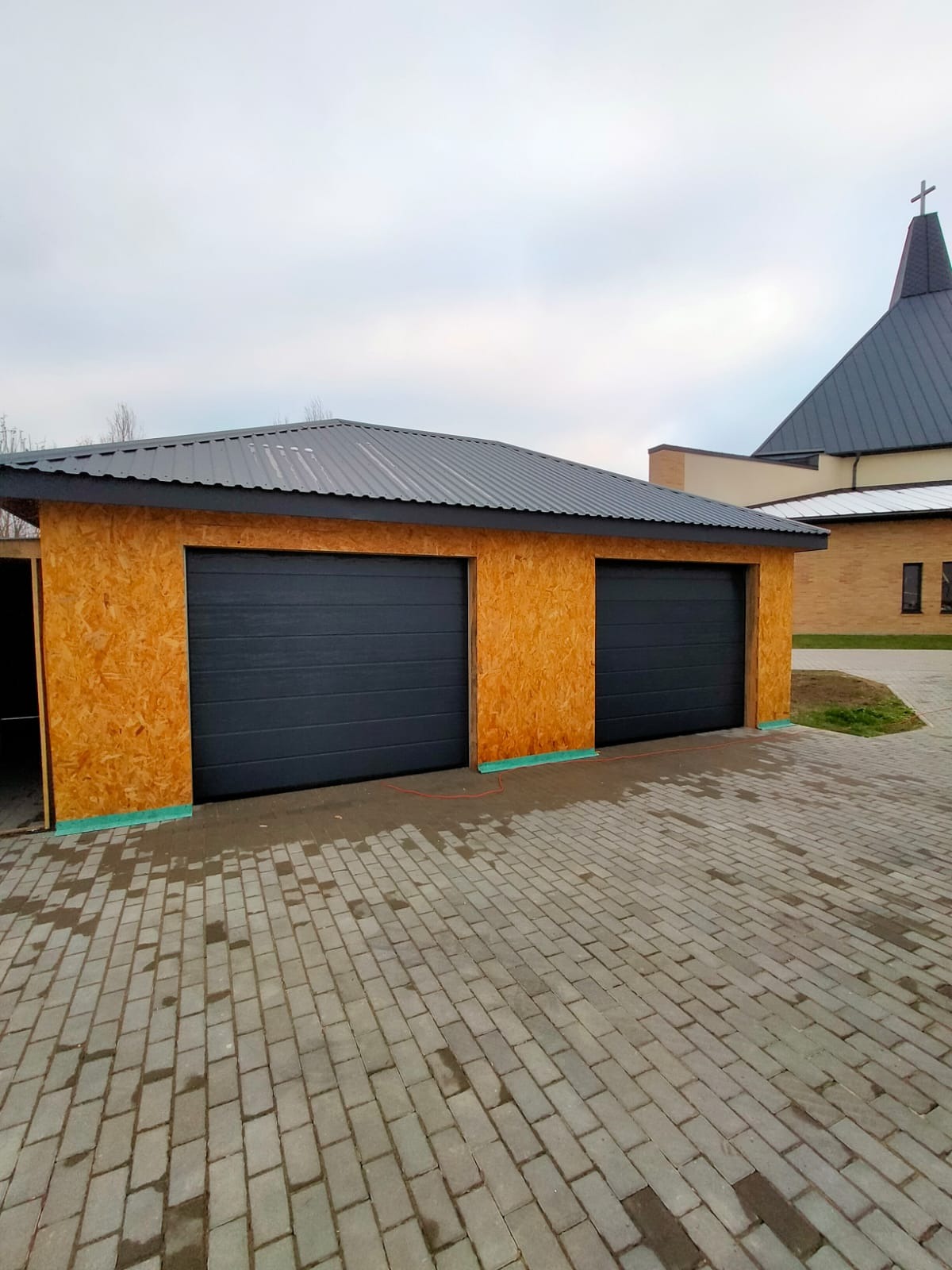 Nowoczesny garaż dwustanowiskowy z szarymi bramami segmentowymi i ścianami z płyty OSB, na tle kościoła z szarą blachą trapezową.