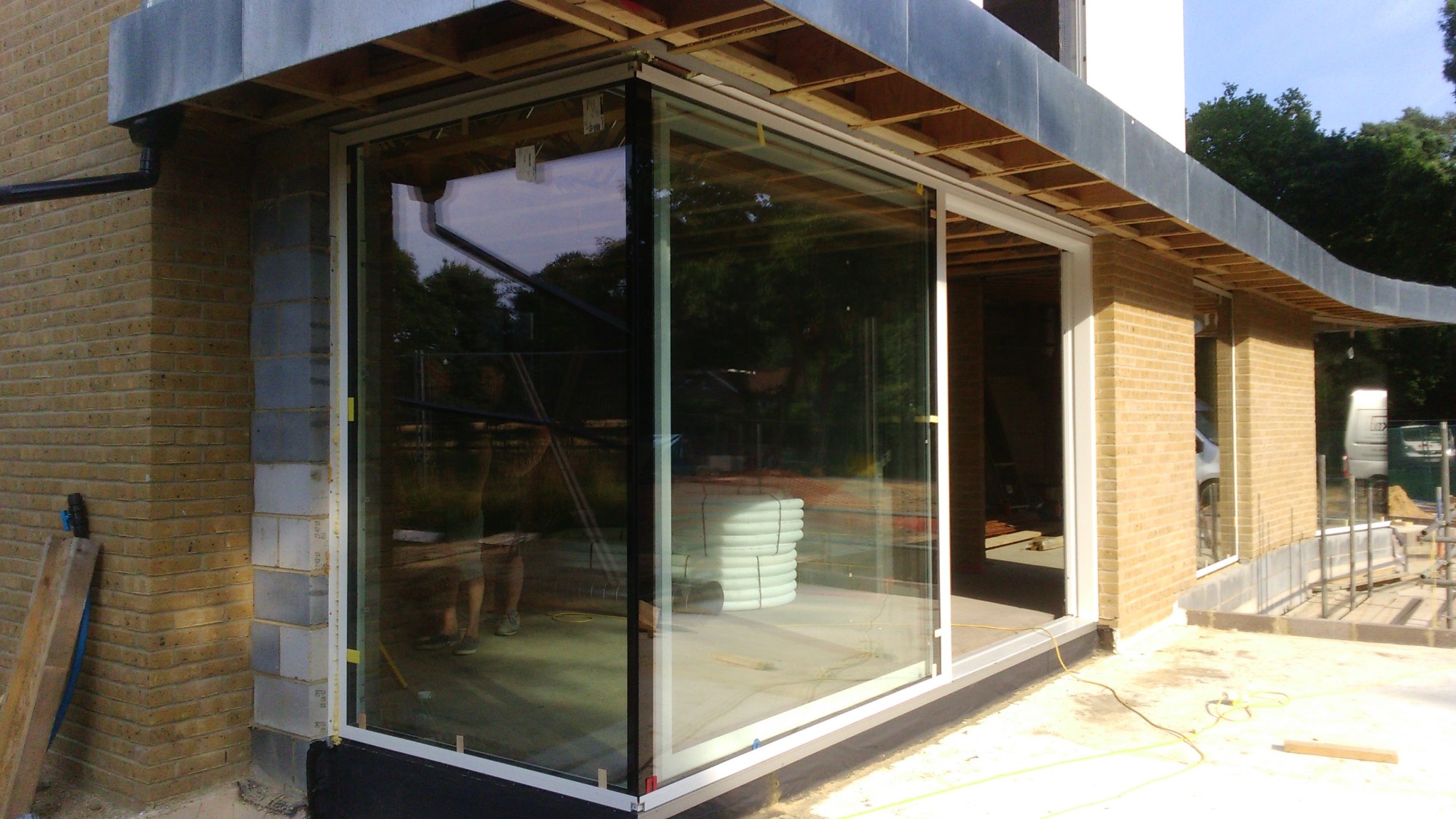 Duże, przesuwne drzwi szklane z białymi ramami w nowym budynku z cegły, widoczne materiały budowlane wewnątrz. Częściowe odbicie osoby w szkle.