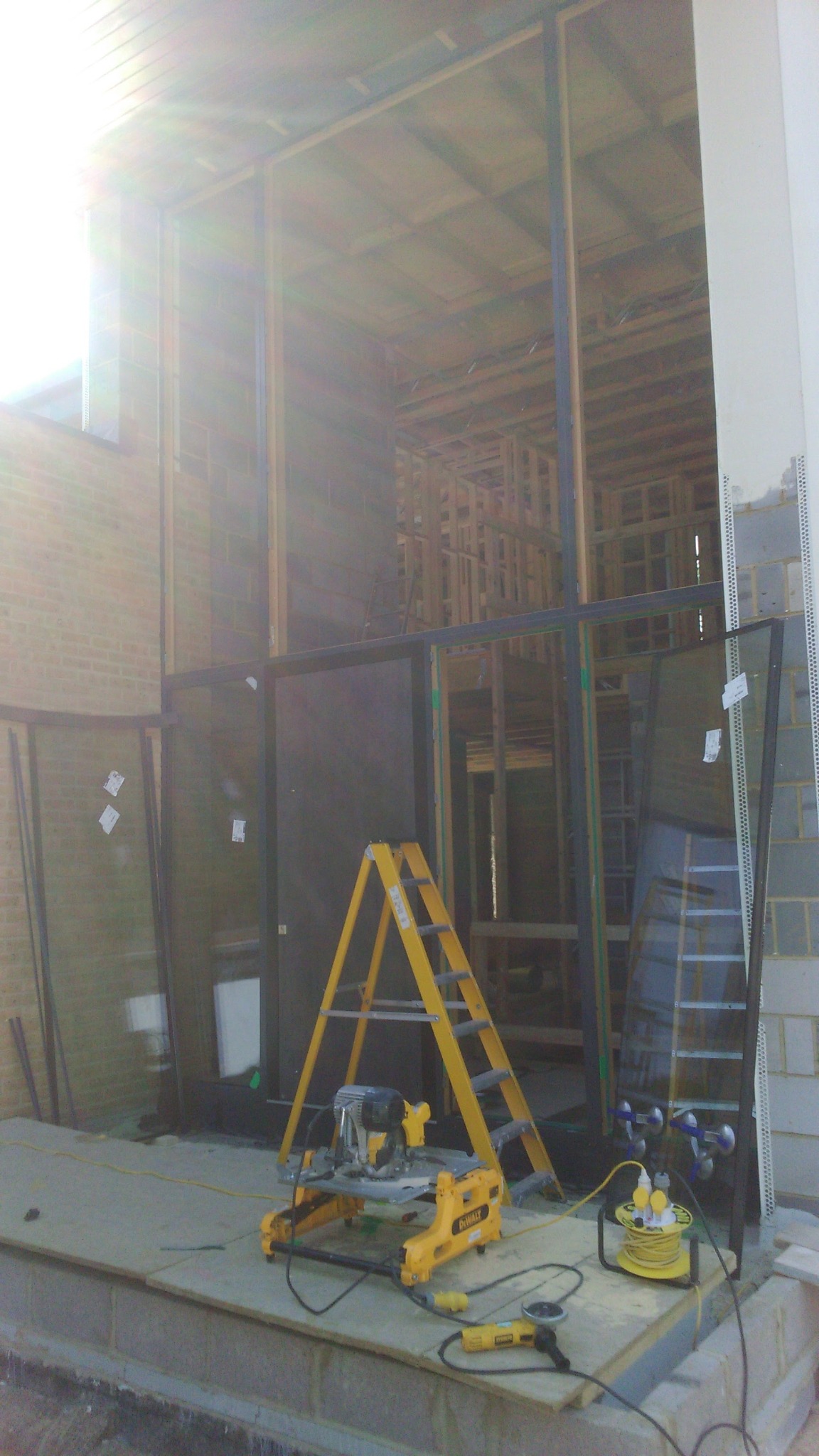 Montaż dużych okien w surowym stanie budynku. Widoczne narzędzia, drabina i ramy okienne. Duże przeszklenia w trakcie instalacji.