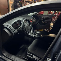 Piotr Dziubek - Wnętrze samochodu Opel Insignia z widocznymi czarnymi fotelami, deską rozdzielczą i kierownicą. W tle garaż z rowerem i regałami.