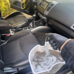 Piotr Dziubek - Wnętrze samochodu z czarną tapicerką, wiadro z brudną wodą w dłoni w czarnej rękawicy, sugerujące czyszczenie tapicerki samochodowej.