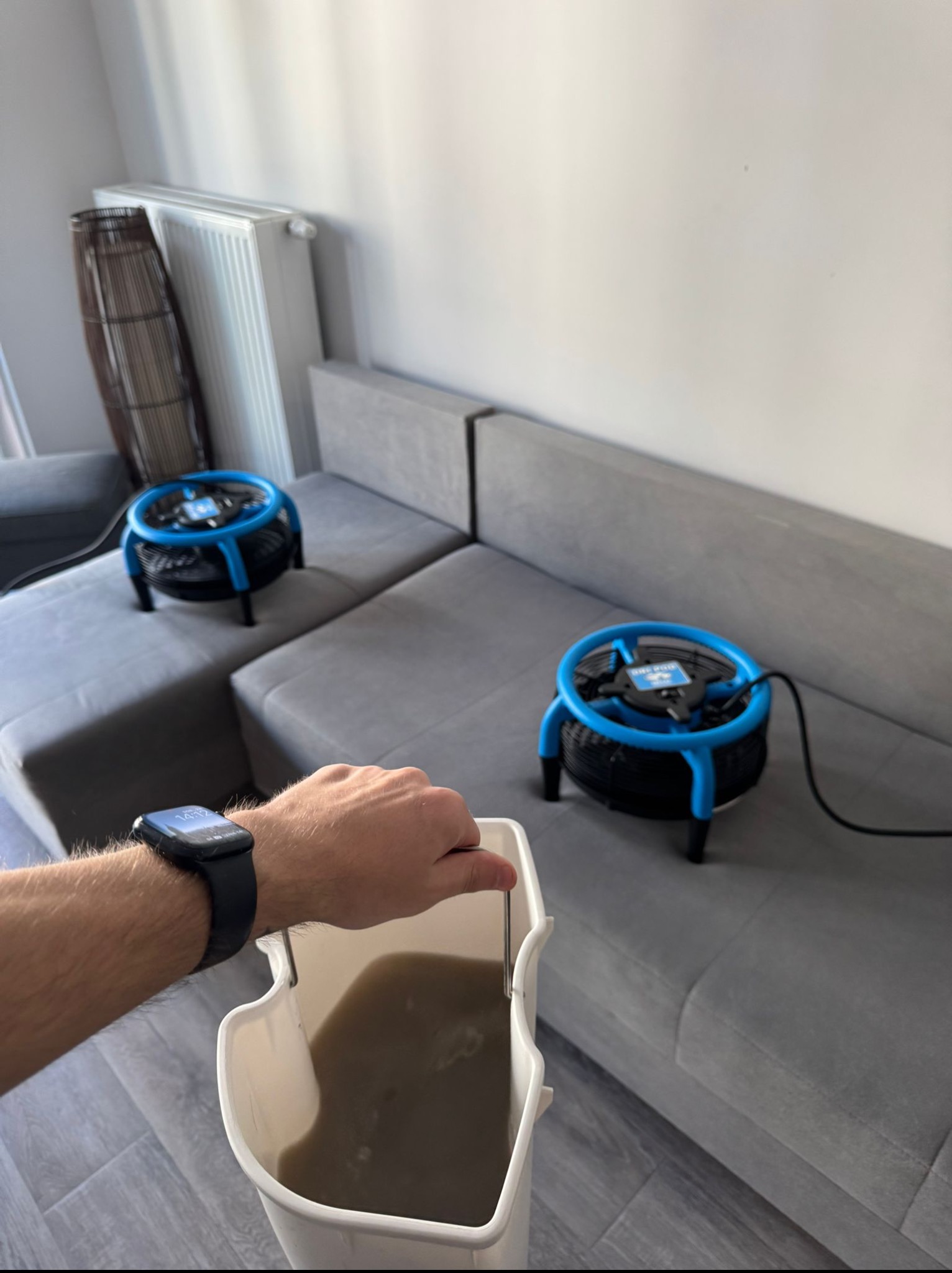 Przenośny wentylator do osuszania tapicerki kanapy po praniu. W tle szara kanapa modułowa i wiadro z brudną wodą. Widoczny zegarek na ręce.