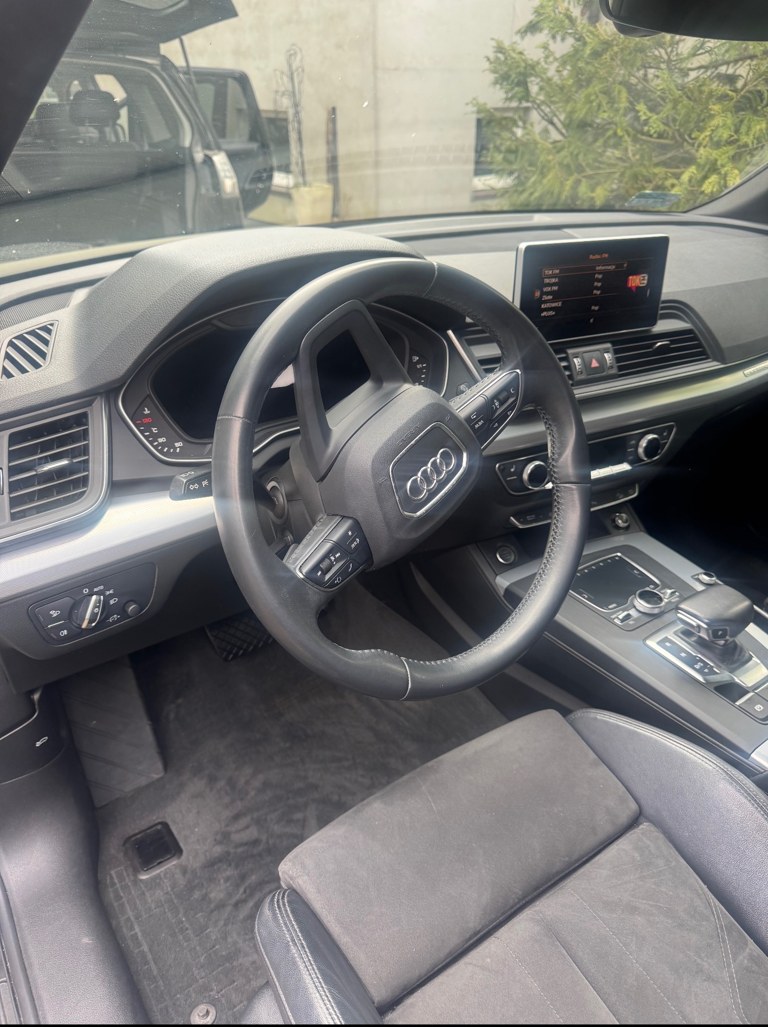 Wnętrze czarnego Audi Q5 z widoczną kierownicą, deską rozdzielczą i częścią fotela kierowcy. Widoczne radio z ekranem. Na zewnątrz zieleń drzew.