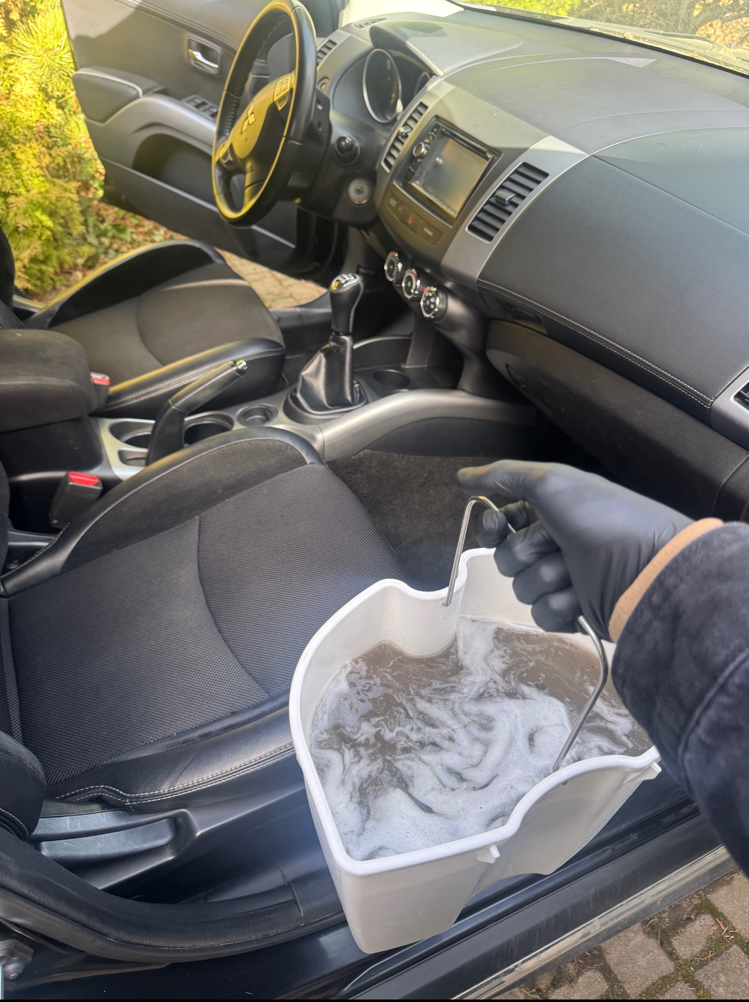 Wnętrze samochodu z czarną tapicerką, wiadro z brudną wodą w dłoni w czarnej rękawicy, sugerujące czyszczenie tapicerki samochodowej.