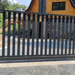 Mariusz Urbanik „Mario” – Profesjonalne Wyroby Spawalnicze - Nowoczesna, przesuwna brama wjazdowa z pionowych profili, w kolorze grafitowym, na tle drewnianego domu i zieleni. Widoczny fragment kostki brukowej.