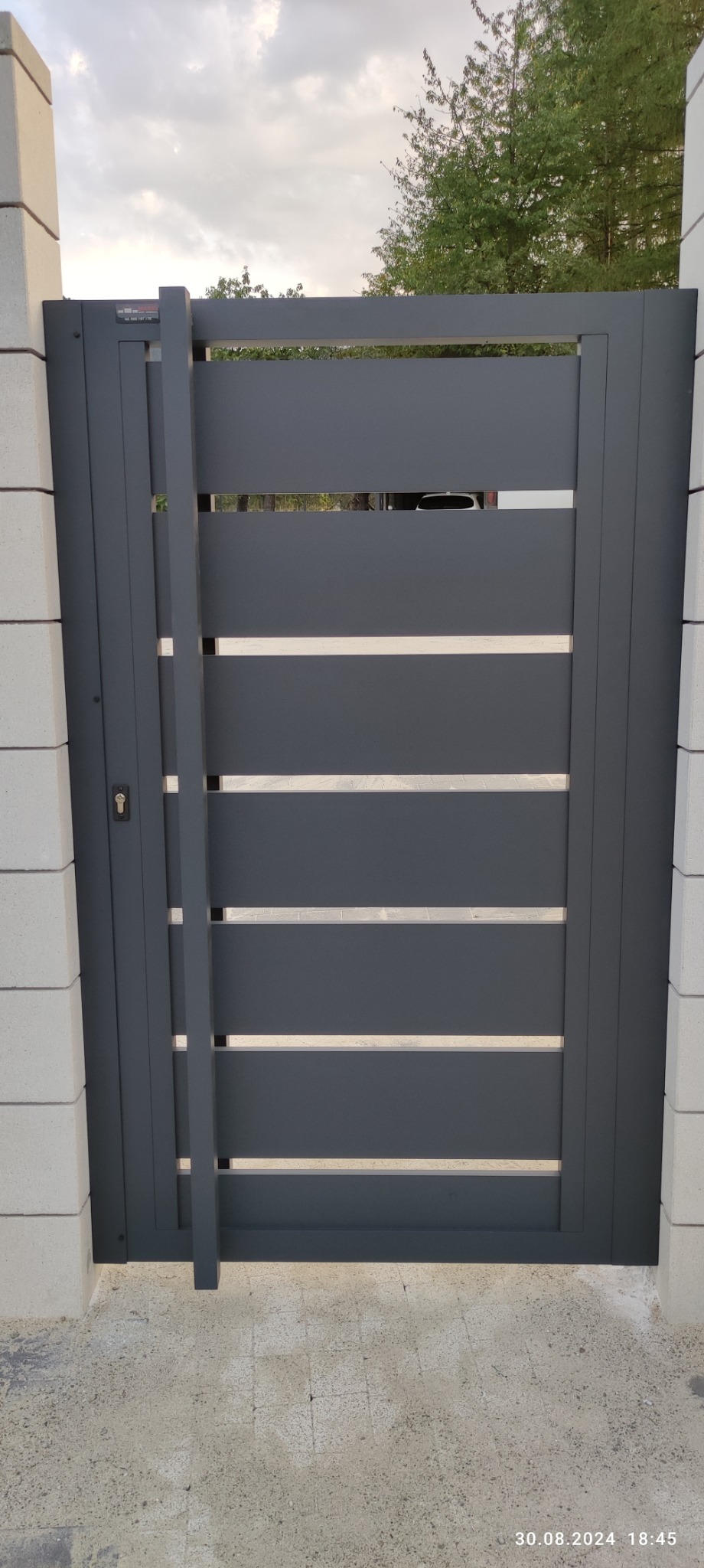 Nowoczesna, grafitowa brama wjazdowa z poziomymi szczeblami, zamontowana między białymi słupkami z bloczków betonowych. Widoczny fragment podjazdu i zieleń w tle.