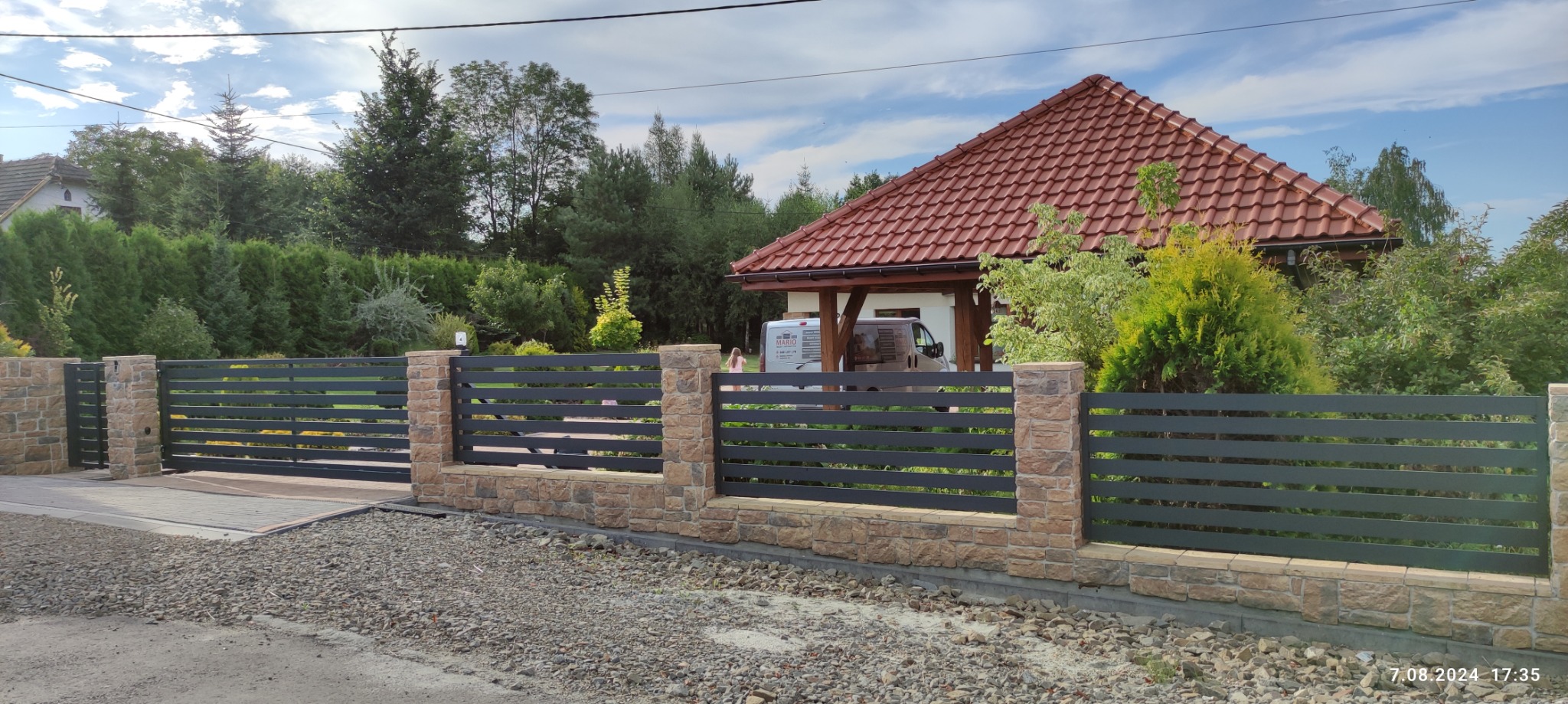 Nowoczesna brama wjazdowa z poziomymi, grafitowymi przęsłami i kamiennymi słupkami na tle zieleni i domu z czerwoną dachówką. Widok z zewnątrz posesji.