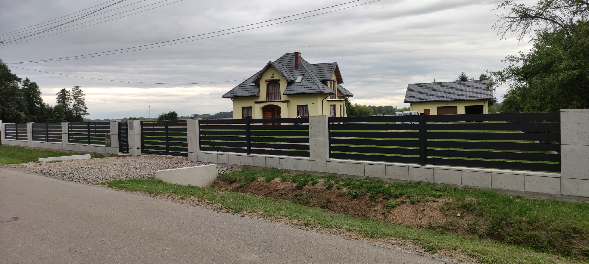 Nowoczesna, pozioma brama wjazdowa i ogrodzenie z czarnych, metalowych paneli na betonowej podmurówce, na tle domu z żółtą elewacją i szarą dachówką.