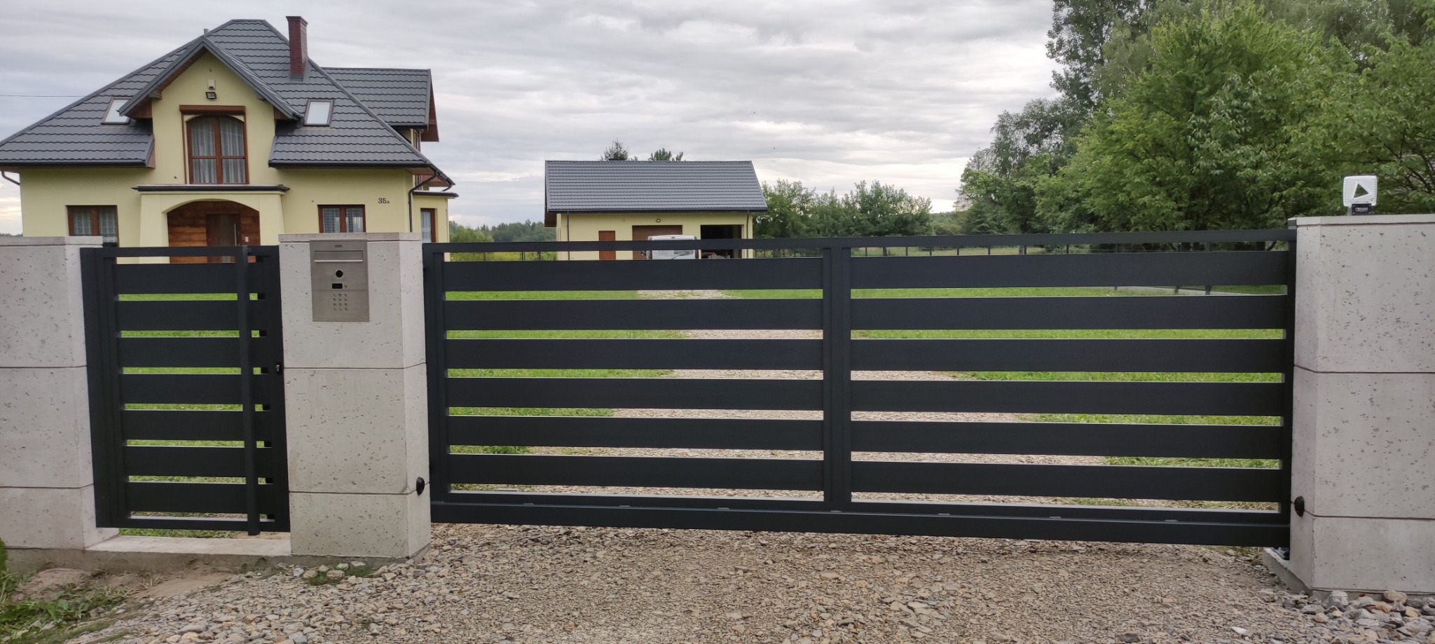 Nowoczesna, pozioma brama wjazdowa w kolorze antracytowym, osadzona na betonowych słupkach z domofonem, na tle domu jednorodzinnego i zieleni.