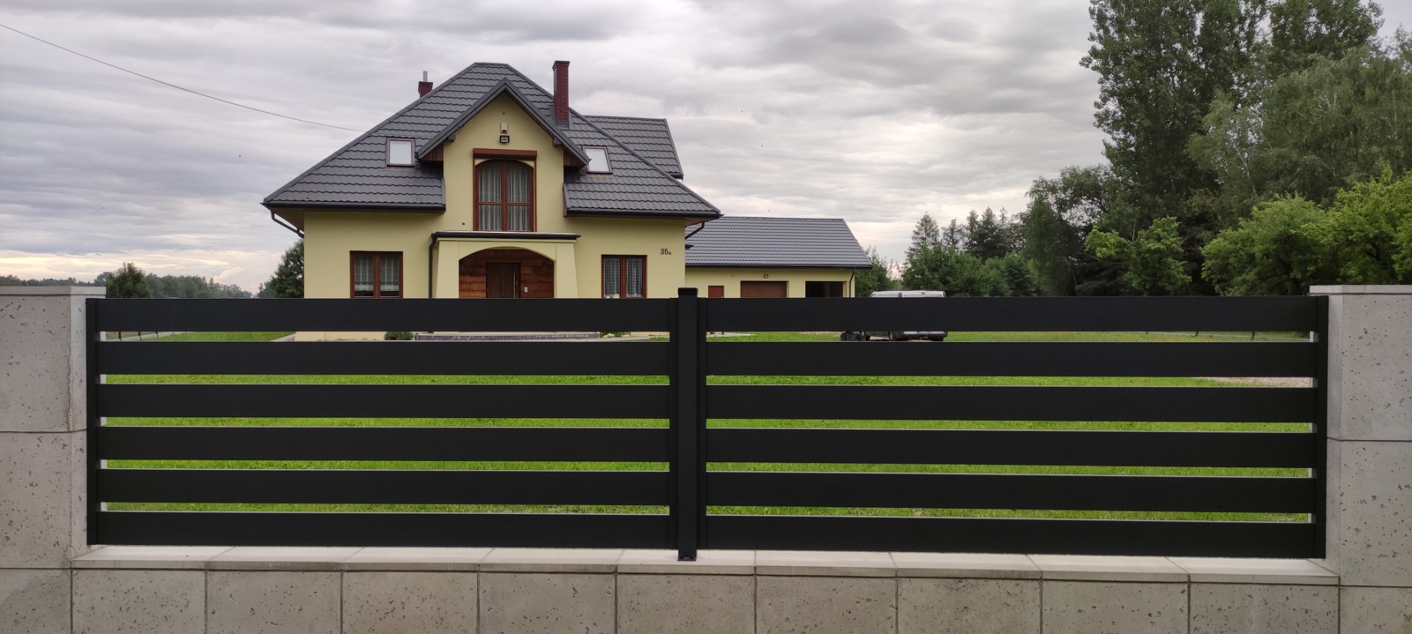 Nowoczesna, pozioma brama wjazdowa z czarnych paneli na tle domu z beżową elewacją i szarą dachówką, otoczona zielenią. Widoczne betonowe słupki i podmurówka.