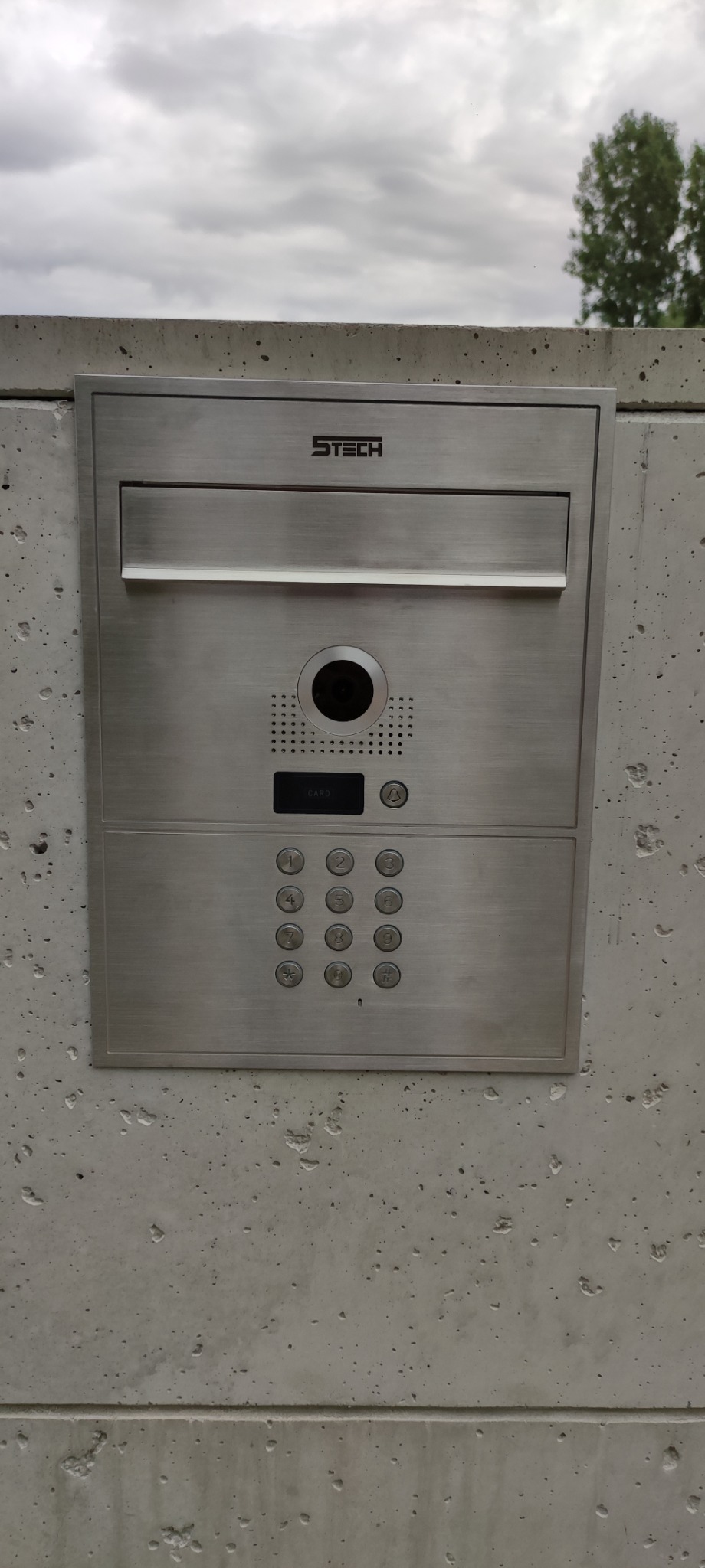 Panel domofonowy STech ze stali nierdzewnej, wbudowany w betonowy mur. Widoczny wizjer, klawiatura numeryczna i czytnik kart. Pochmurne niebo w tle.