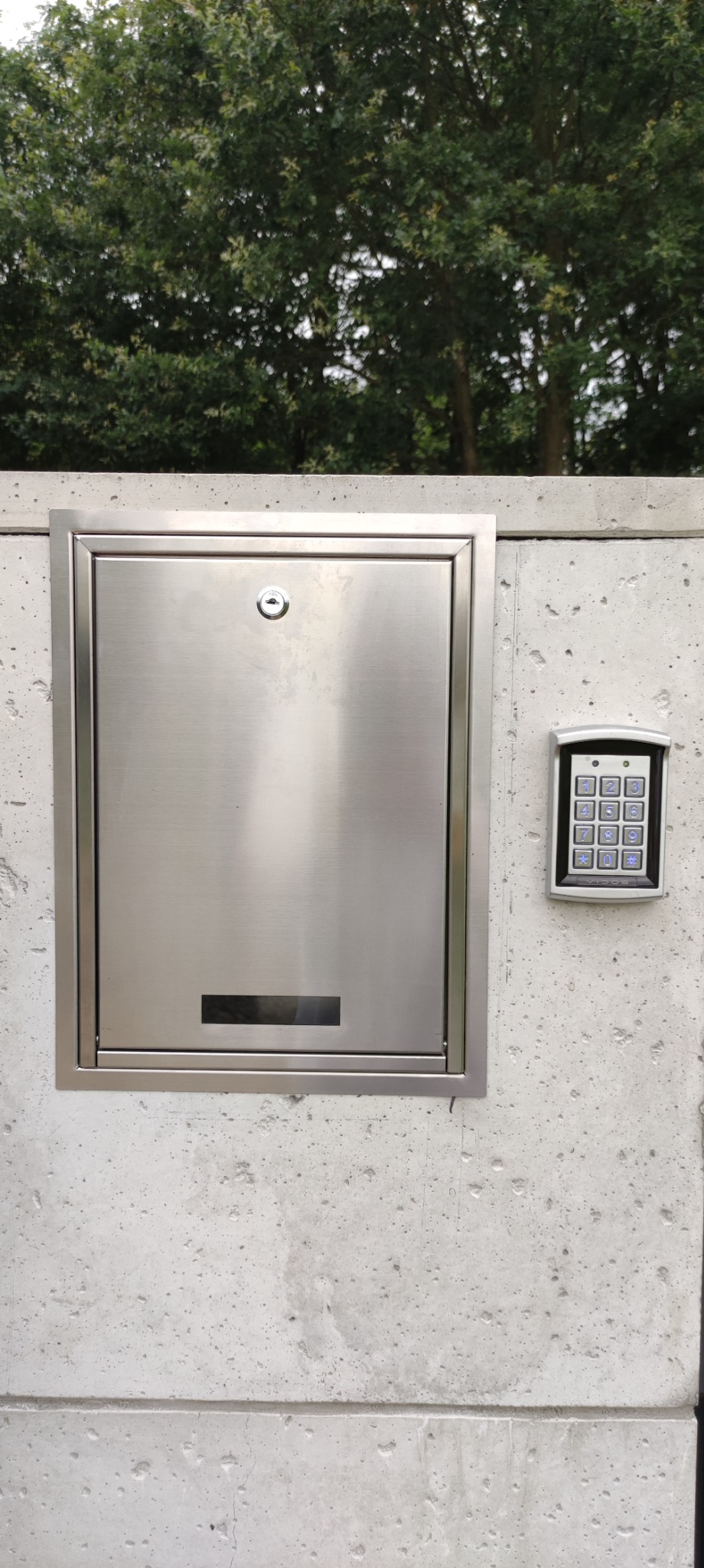 Metalowa skrzynka na listy z zamkiem i panelem numerycznym na betonowym ogrodzeniu, z zielenią w tle. Minimalistyczny design, nowoczesne rozwiązanie.