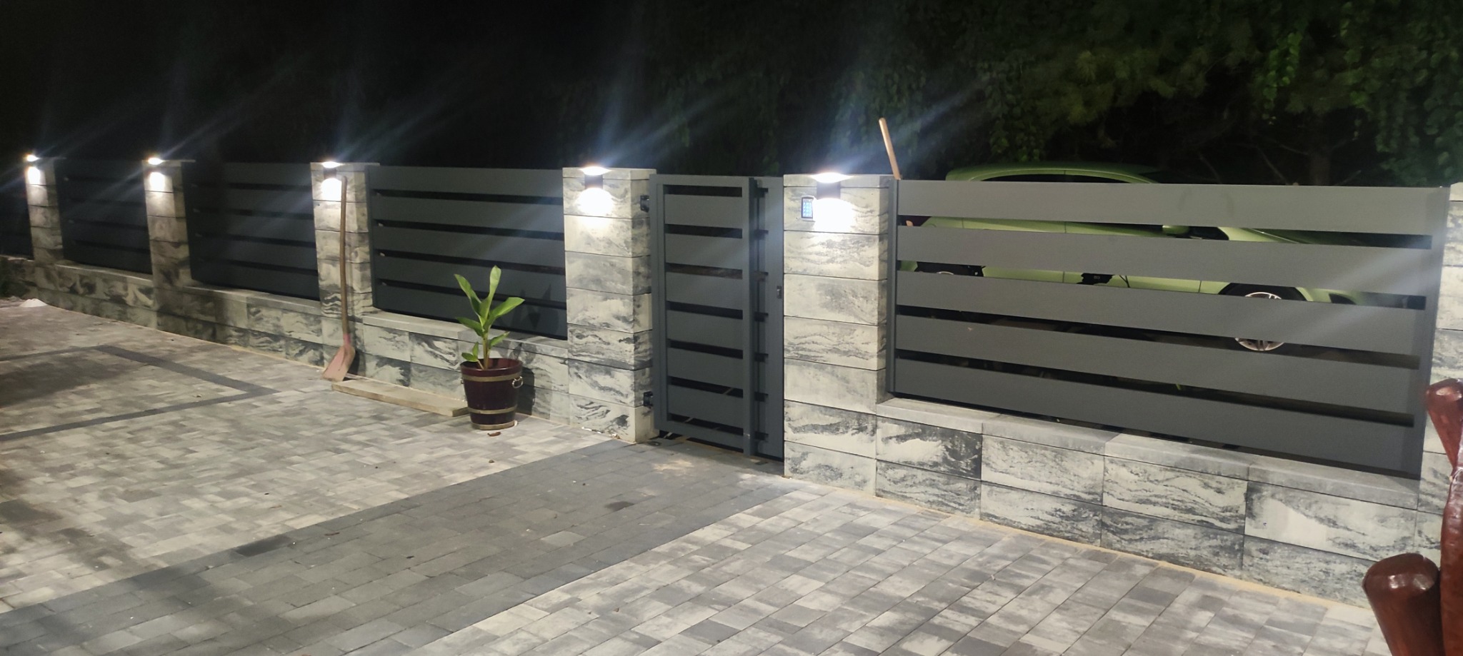 Nowoczesna, szara brama wjazdowa z poziomymi panelami i kamiennymi słupkami oświetlonymi lampami. Widoczny fragment zielonego samochodu za bramą i donica z rośliną.