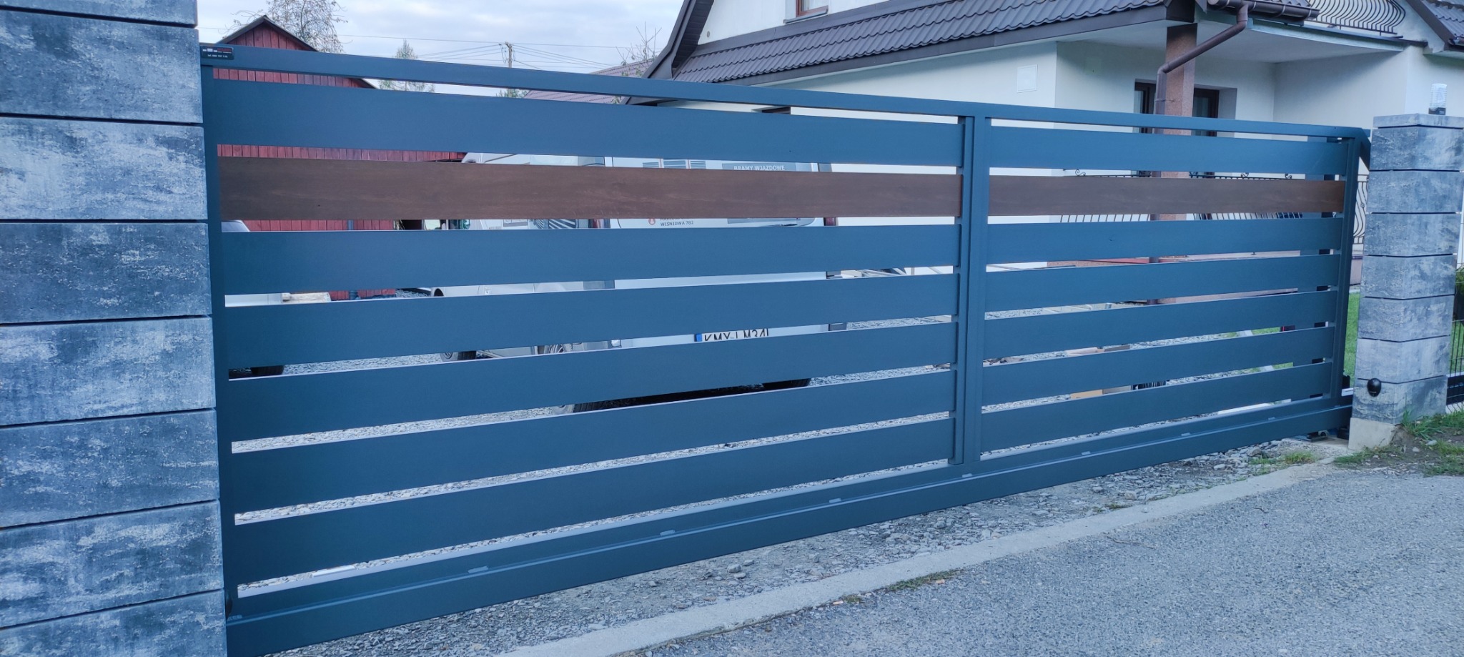 Nowoczesna brama wjazdowa przesuwna, szara, z poziomymi panelami i drewnianym akcentem, osadzona na betonowych słupkach. Widok z zewnątrz posesji.