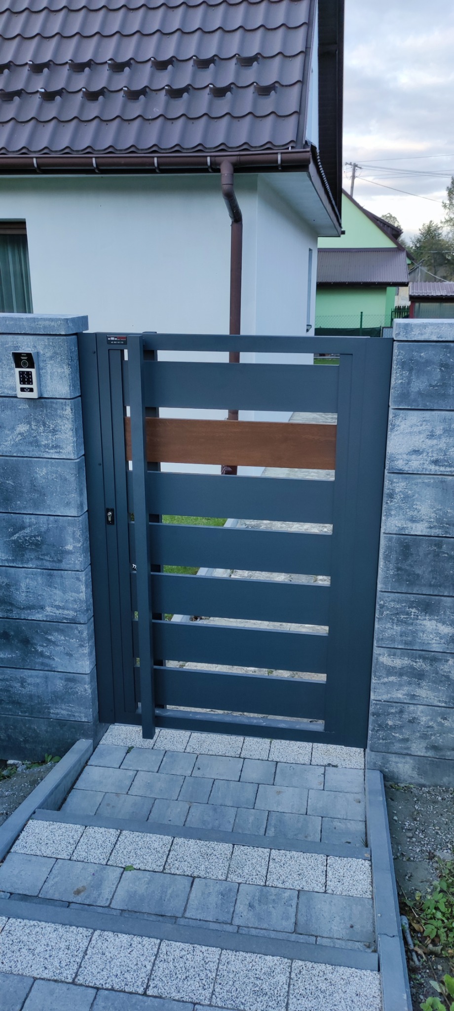 Nowoczesna, grafitowa furtka z drewnianym akcentem, wkomponowana w szare, betonowe słupki. Widoczny wideodomofon i schody z kostki brukowej.