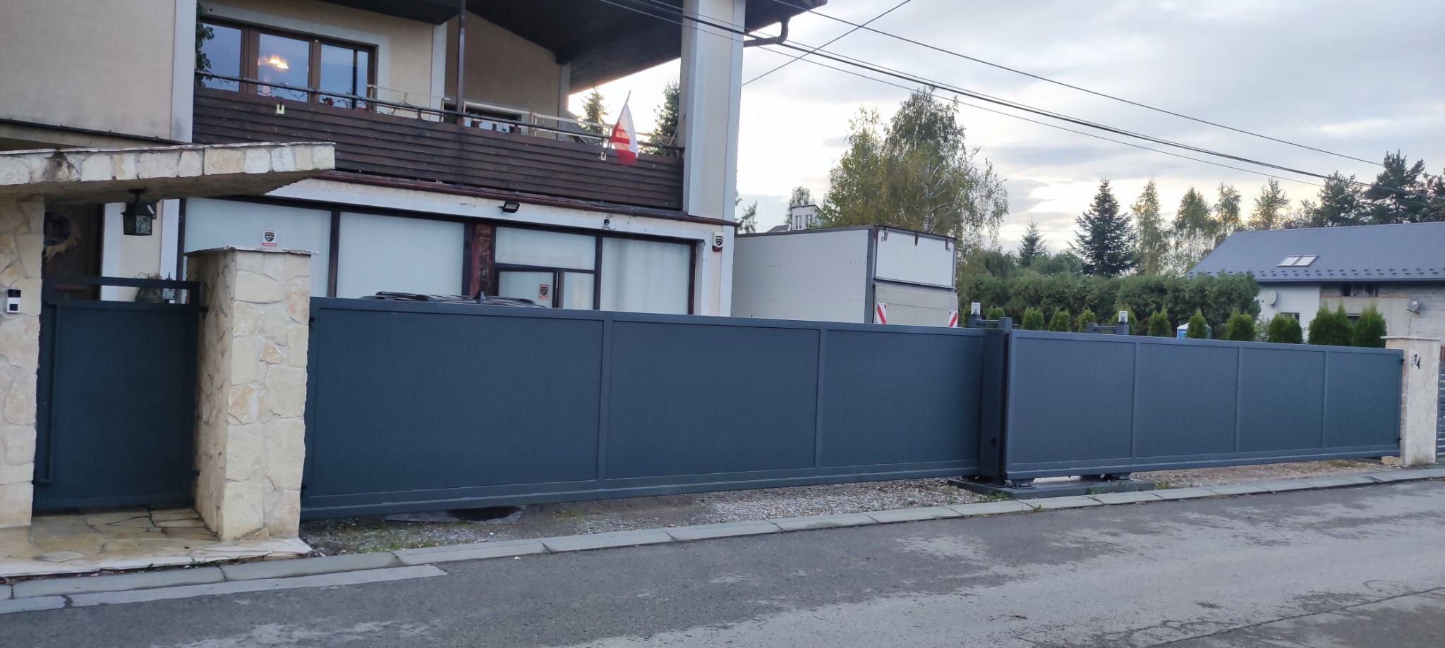 Nowoczesna, przesuwna brama wjazdowa w kolorze grafitowym, z kamiennymi słupkami, na tle domu z balkonem i polską flagą. Widok z poziomu ulicy.