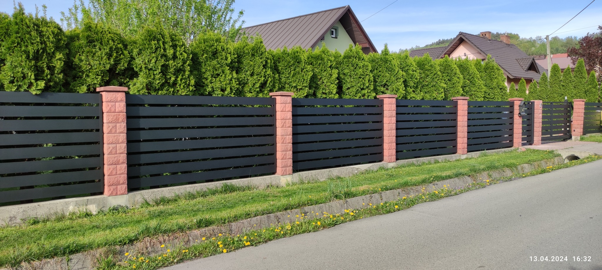 Nowoczesne ogrodzenie panelowe z czarnymi, poziomymi przęsłami i ceglanymi słupkami, otoczone zielenią, z widoczną furtką w tle. Słoneczny dzień.