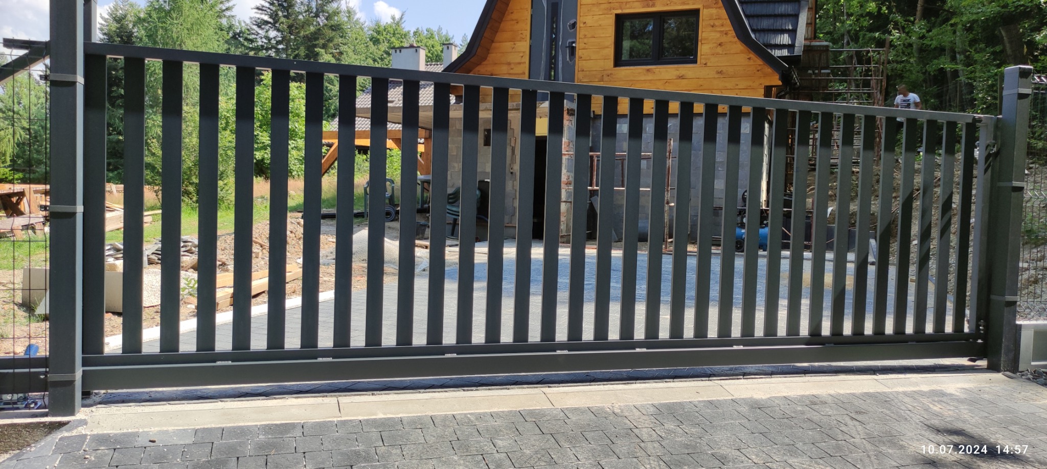 Nowoczesna, przesuwna brama wjazdowa z pionowych profili, w kolorze grafitowym, na tle drewnianego domu i zieleni. Widoczny fragment kostki brukowej.