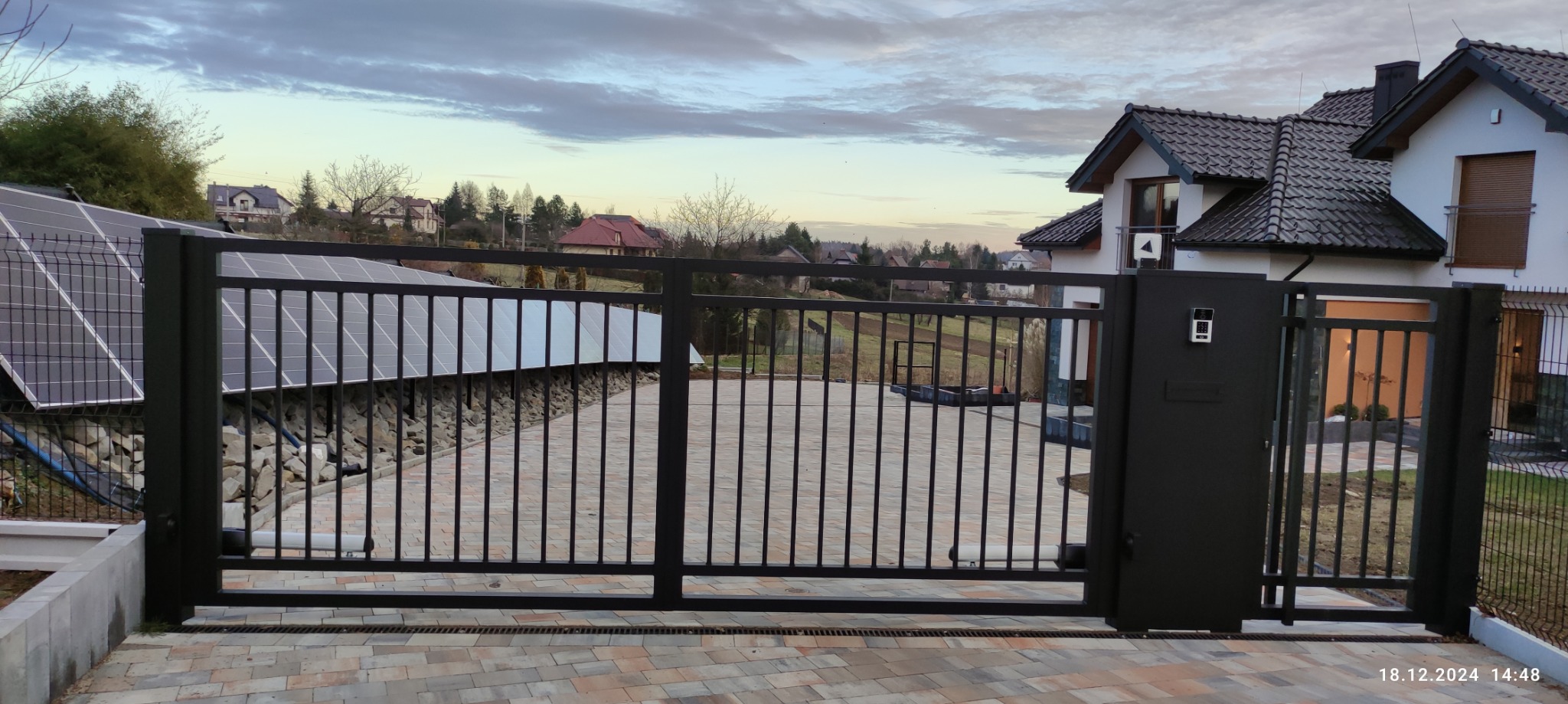 Nowoczesna, przesuwna brama wjazdowa w kolorze antracytowym, z panelem fotowoltaicznym w tle. Widoczny dom jednorodzinny i kostka brukowa na podjeździe.