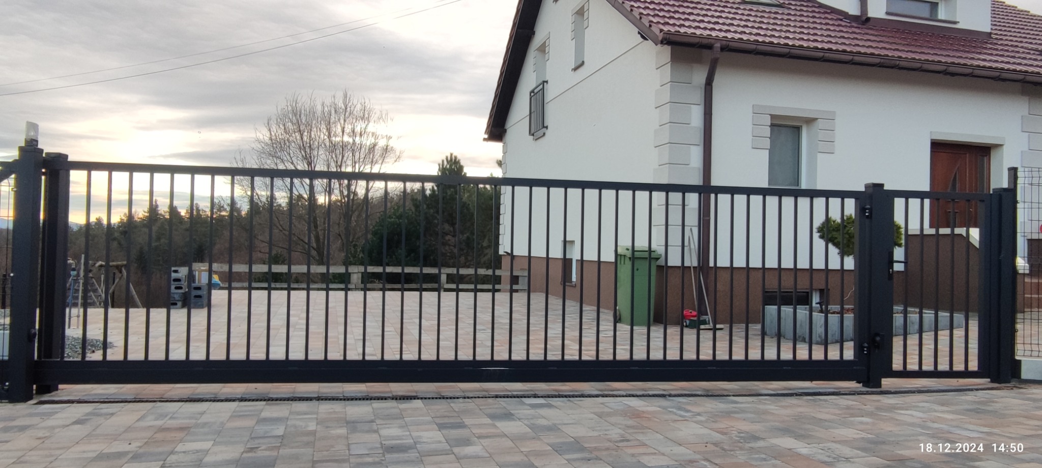 Nowoczesna, czarna brama wjazdowa z pionowymi szczebelkami na tle domu z beżową elewacją i kostki brukowej. Widoczne elementy automatyki.