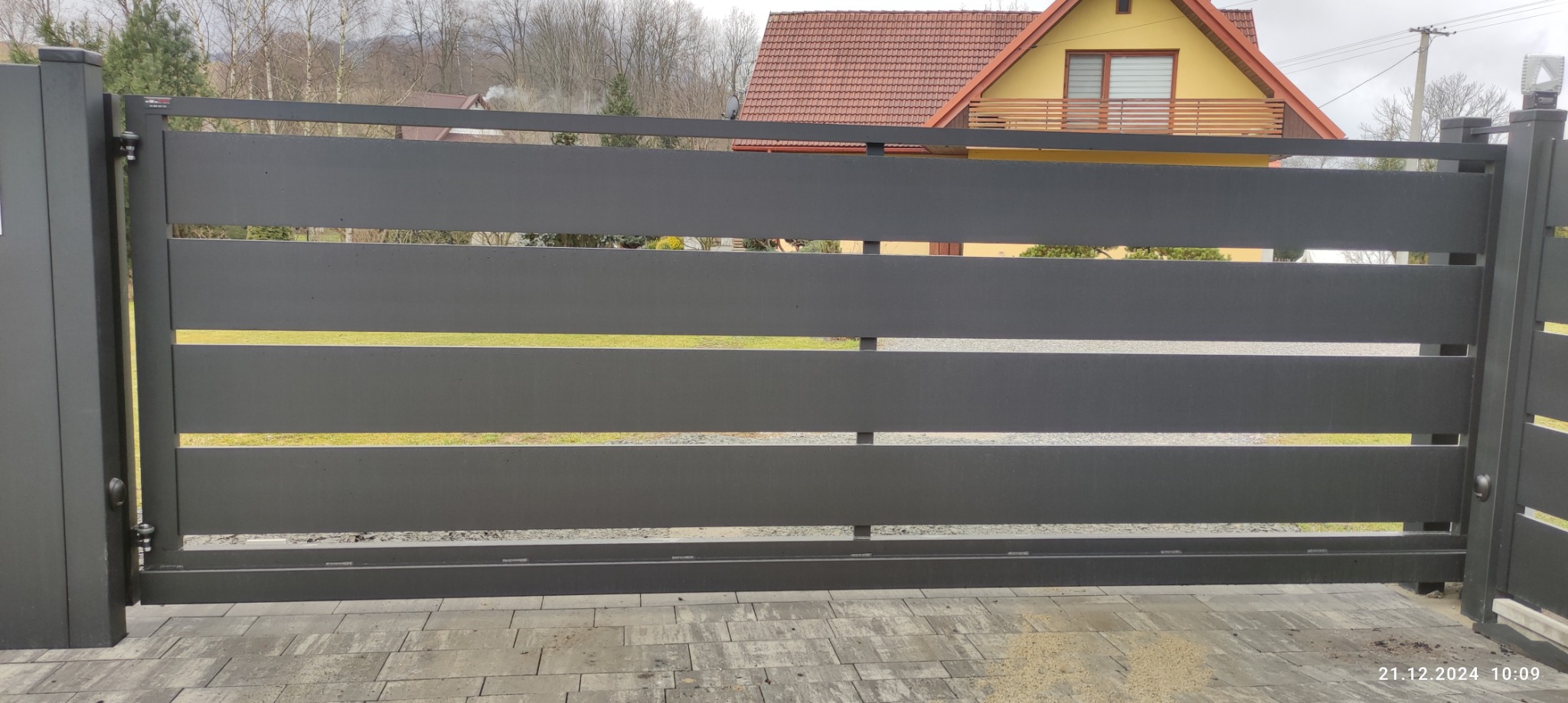 Nowoczesna, pozioma brama wjazdowa w kolorze antracytowym, z widocznym domem w tle. Minimalistyczny design, poziome panele z prześwitami, kostka brukowa u dołu.