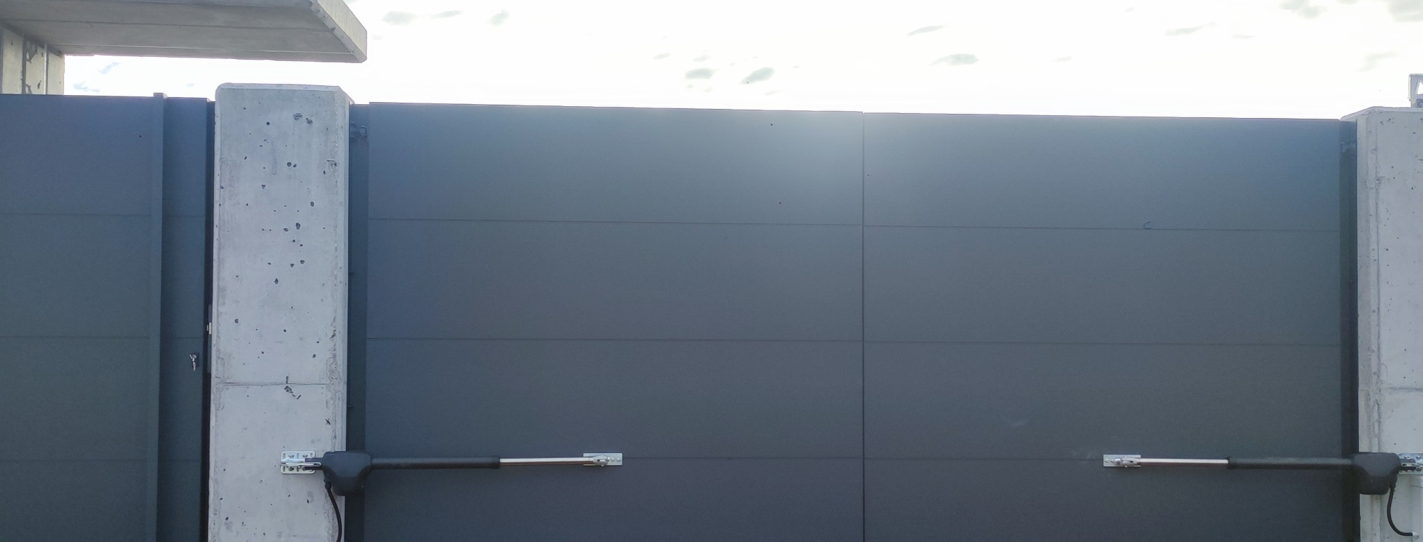 Nowoczesna, przesuwna brama wjazdowa w kolorze grafitowym z automatyką, zamontowana między betonowymi słupkami. Widoczne siłowniki bramy.