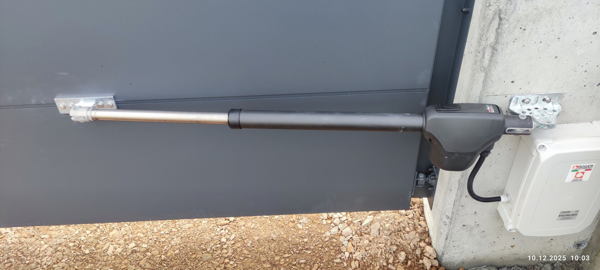 Szary napęd ramieniowy do bramy wjazdowej, zamontowany na betonowym słupku. Widoczna skrzynka sterownicza z logo Roger Technology.