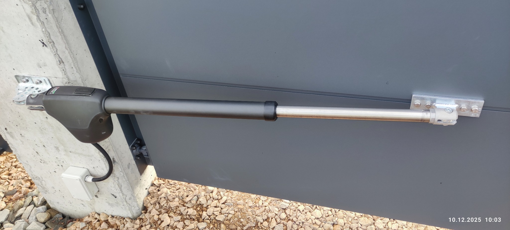 Zamontowany siłownik bramy wjazdowej, widoczne mocowanie do betonowego słupka i skrzydła bramy. Szara brama, jasny słupek i żwir.