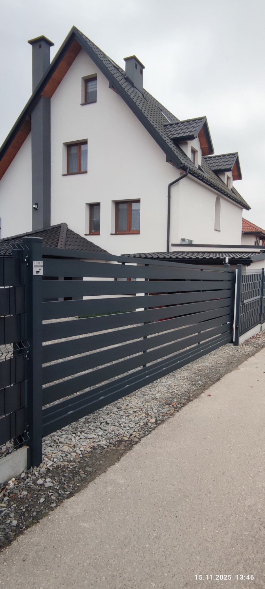 Nowoczesna, pozioma brama wjazdowa w kolorze grafitowym, z widocznym fragmentem domu w tle. Brama zamontowana na posesji, widoczne kamienie ozdobne.