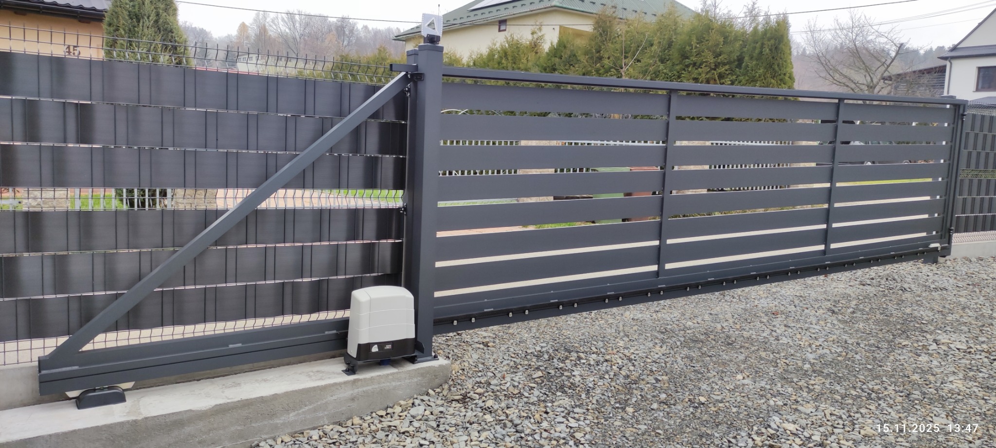 Nowoczesna, przesuwna brama wjazdowa w kolorze grafitowym z poziomymi szczeblami i automatyką, zamontowana na betonowej podstawie. Widok z zewnątrz posesji.