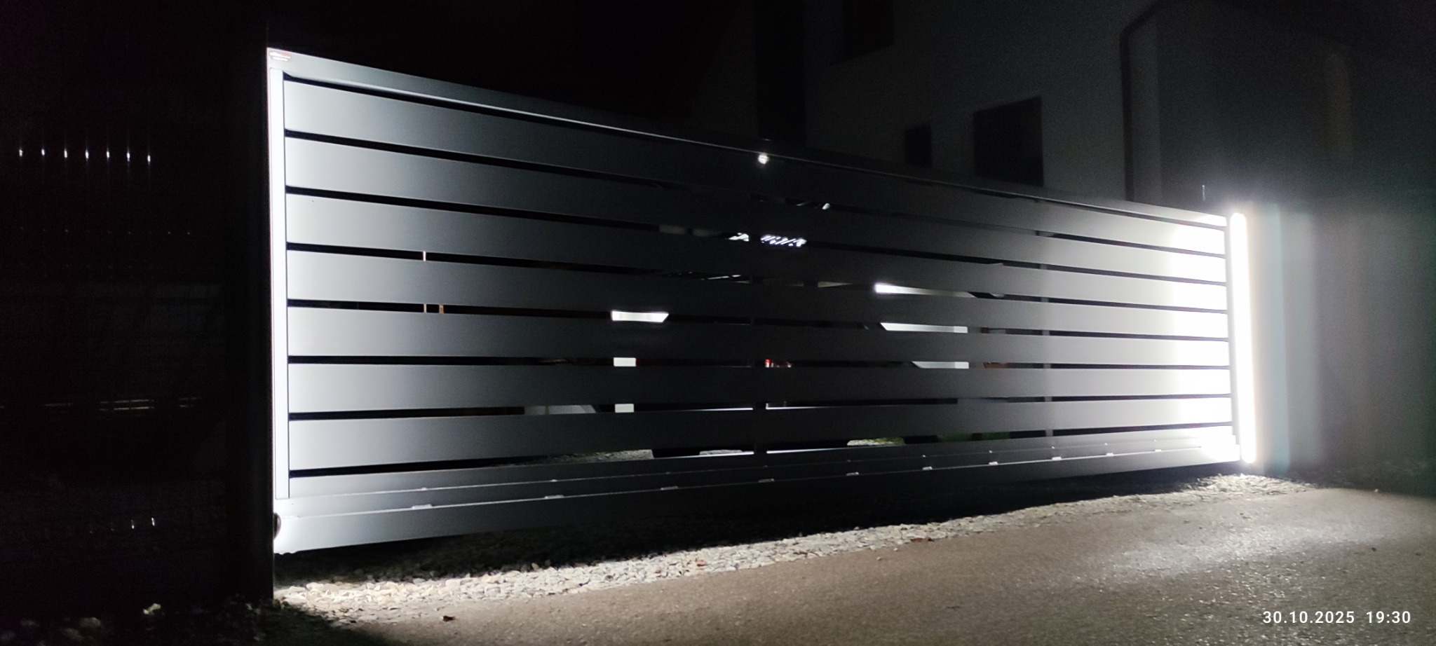 Nowoczesna brama wjazdowa z poziomych paneli, oświetlona od dołu, widoczna nocą. Minimalistyczny design, szara kolorystyka, subtelne światło podkreśla detale.