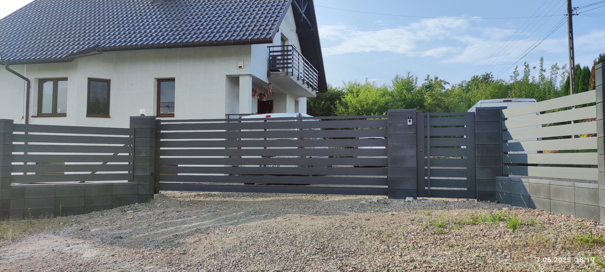 Nowoczesna brama wjazdowa z poziomych, szarych profili, osadzona na betonowych słupkach. W tle biały dom z balkonem i zielone drzewa. Widok z zewnątrz posesji.