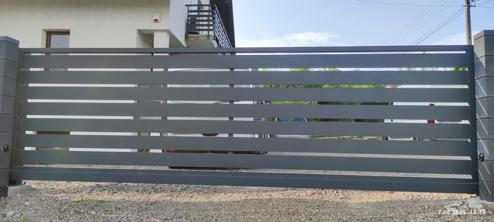 Nowoczesna, przesuwna brama wjazdowa w kolorze grafitowym z poziomymi panelami, na tle jasnego domu i podjazdu z kamienia. Widoczne słupki z szarej kostki.