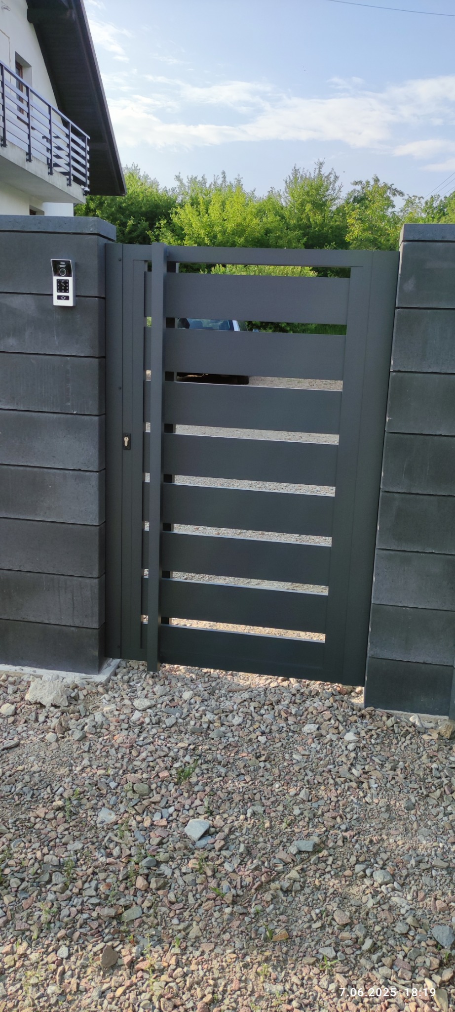 Nowoczesna, grafitowa brama wjazdowa z poziomymi szczeblami, zamontowana między słupkami z szarego betonu architektonicznego. Widoczny dom i podjazd.